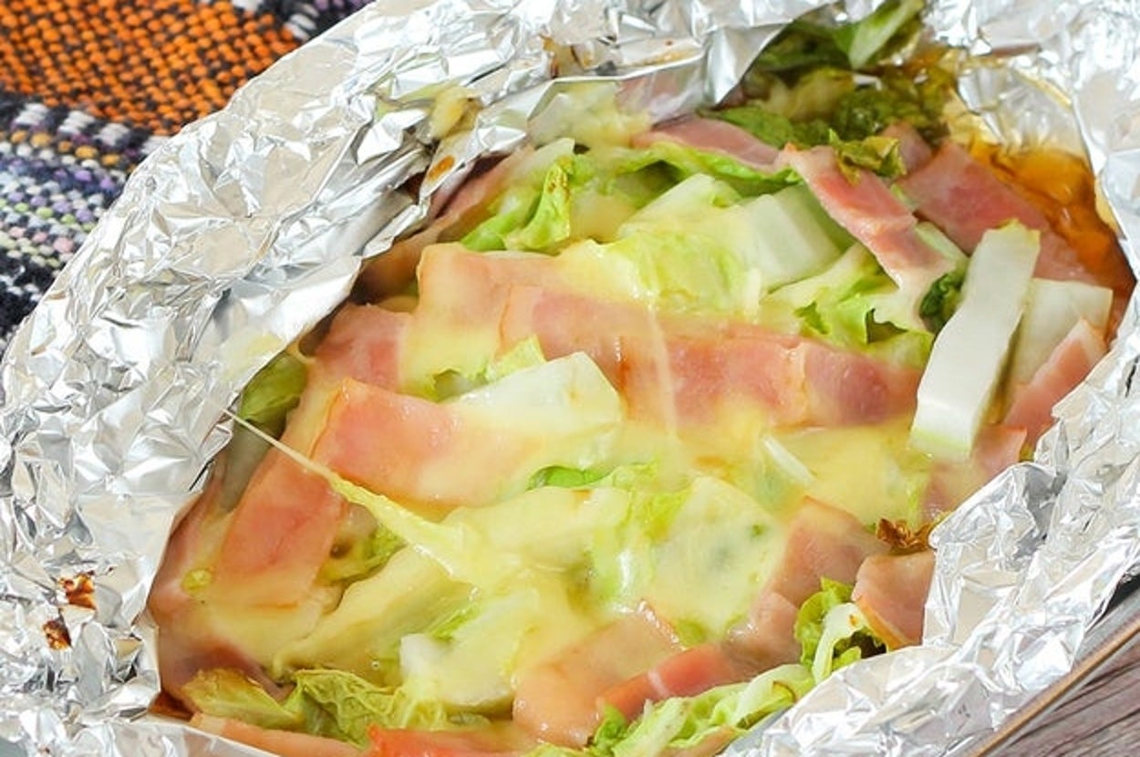 アルミホイルに包まれたキャベツとベーコンの上にチーズがかかった料理。背景に布がある。