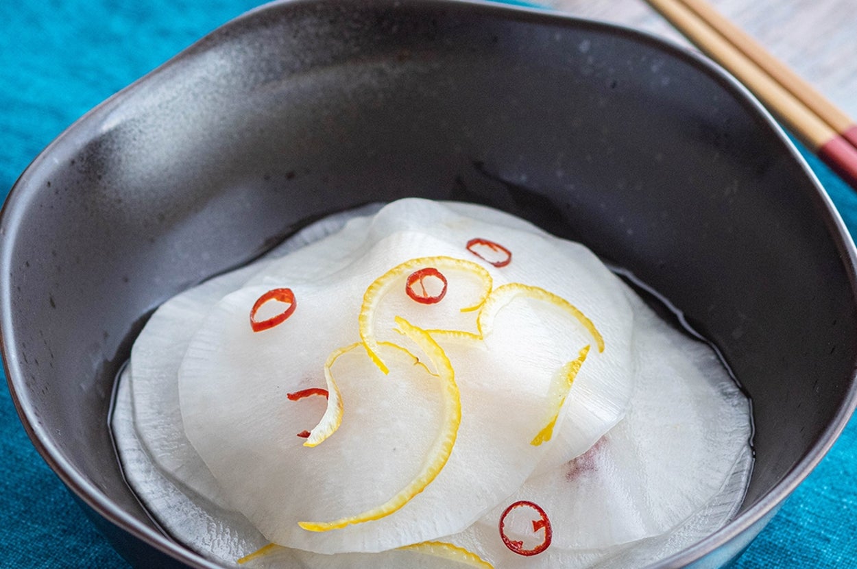 薄くスライスされた大根が黒い器に盛られた料理。上に赤と黄色の調味料で飾り付け。背景に箸と飲み物。