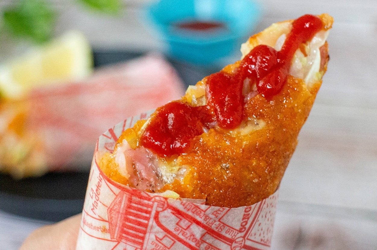 手に持たれたホットドッグが紙に包まれており、上にはケチャップがかかっています。背景にソースとレモンの一片があります。