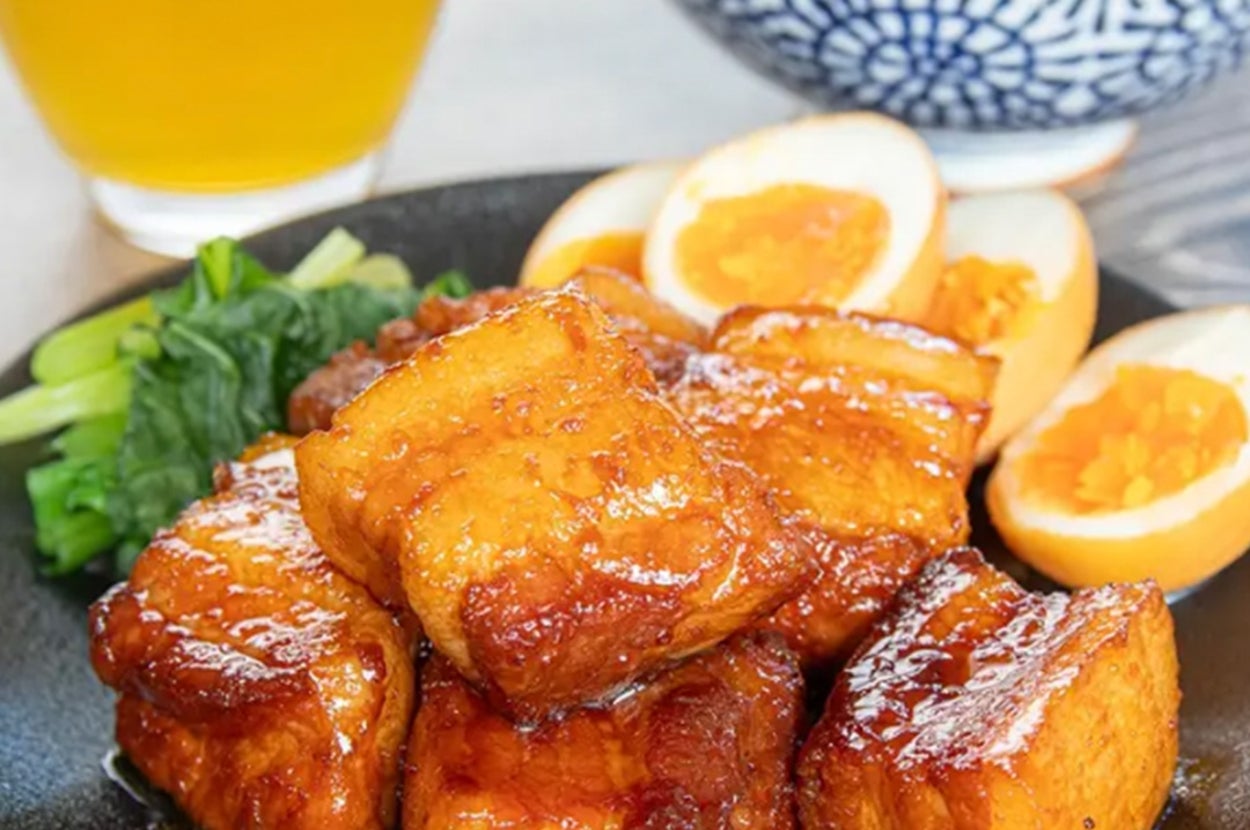 こんがり焼かれた角煮が皿に盛られ、半熟卵と青菜が添えられた料理。後ろにご飯と飲み物が見える。