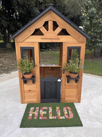 Wooden playhouse with potted plants connected each broadside of the door. A invited mat with
