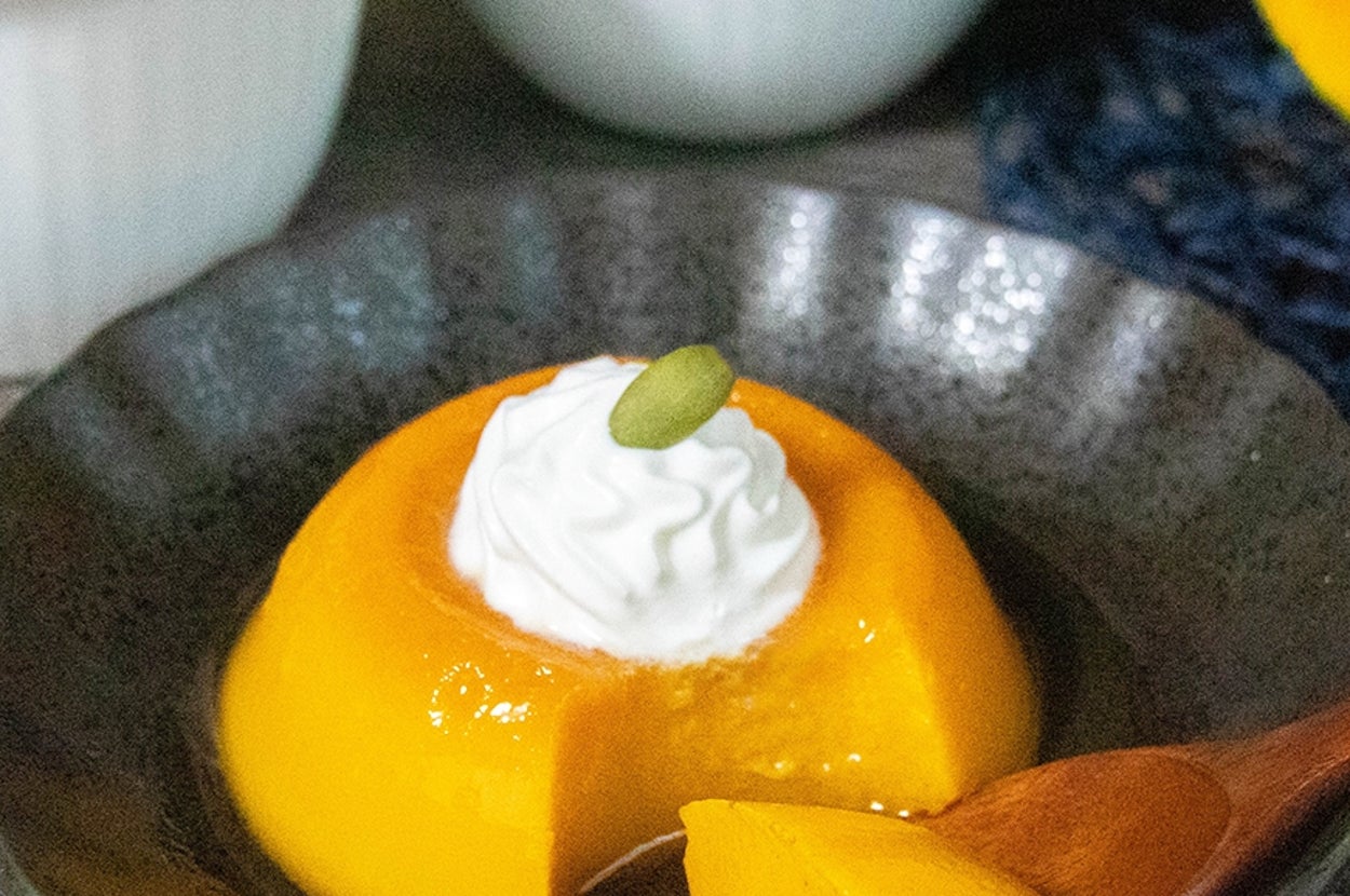 かぼちゃのプリンが黒い皿に盛られ、クリームと種がトッピングされています。背景に白いカップが2つあります。