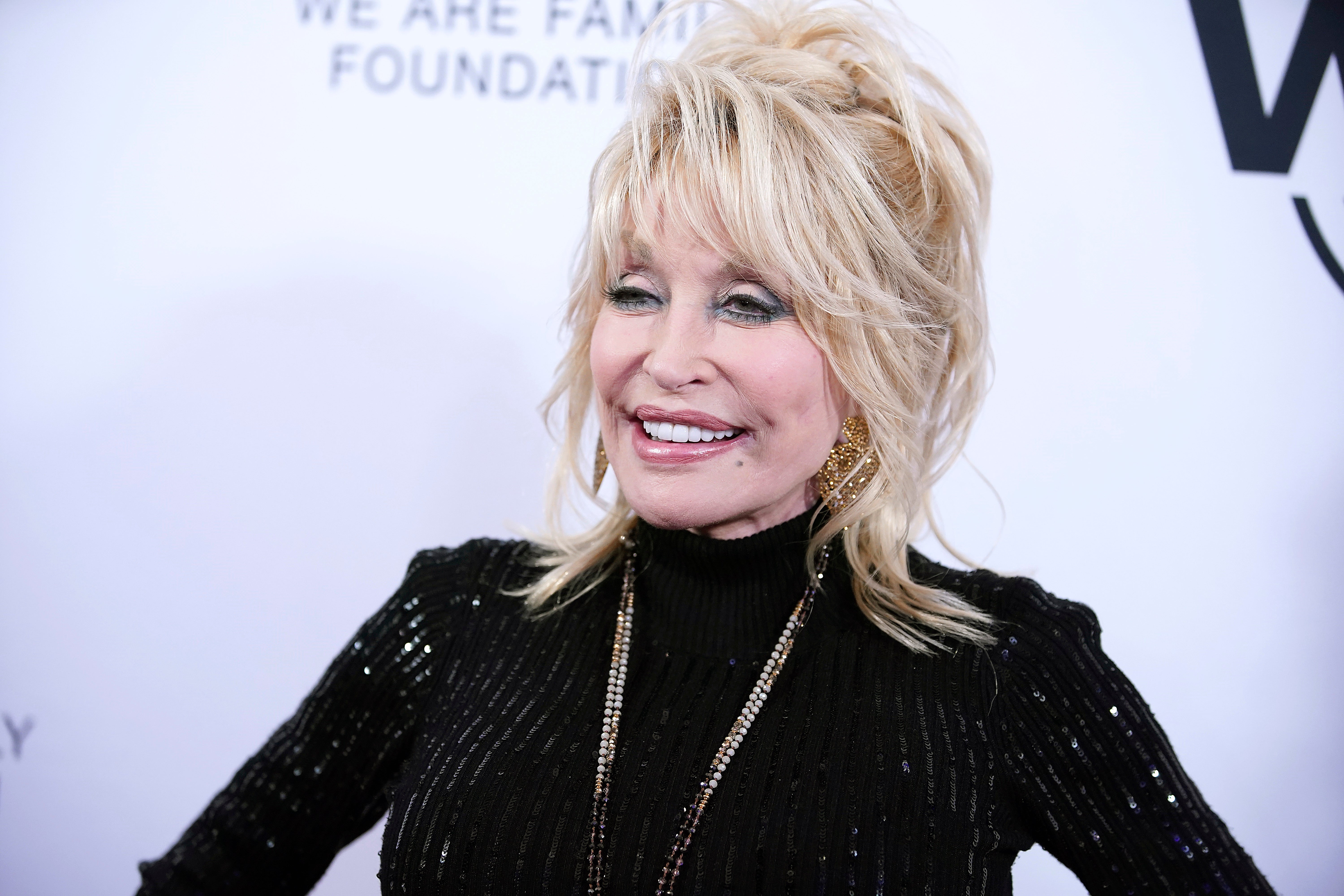 Person in a shimmering black dress with sequins and layered blonde hair, smiling on the red carpet at an event