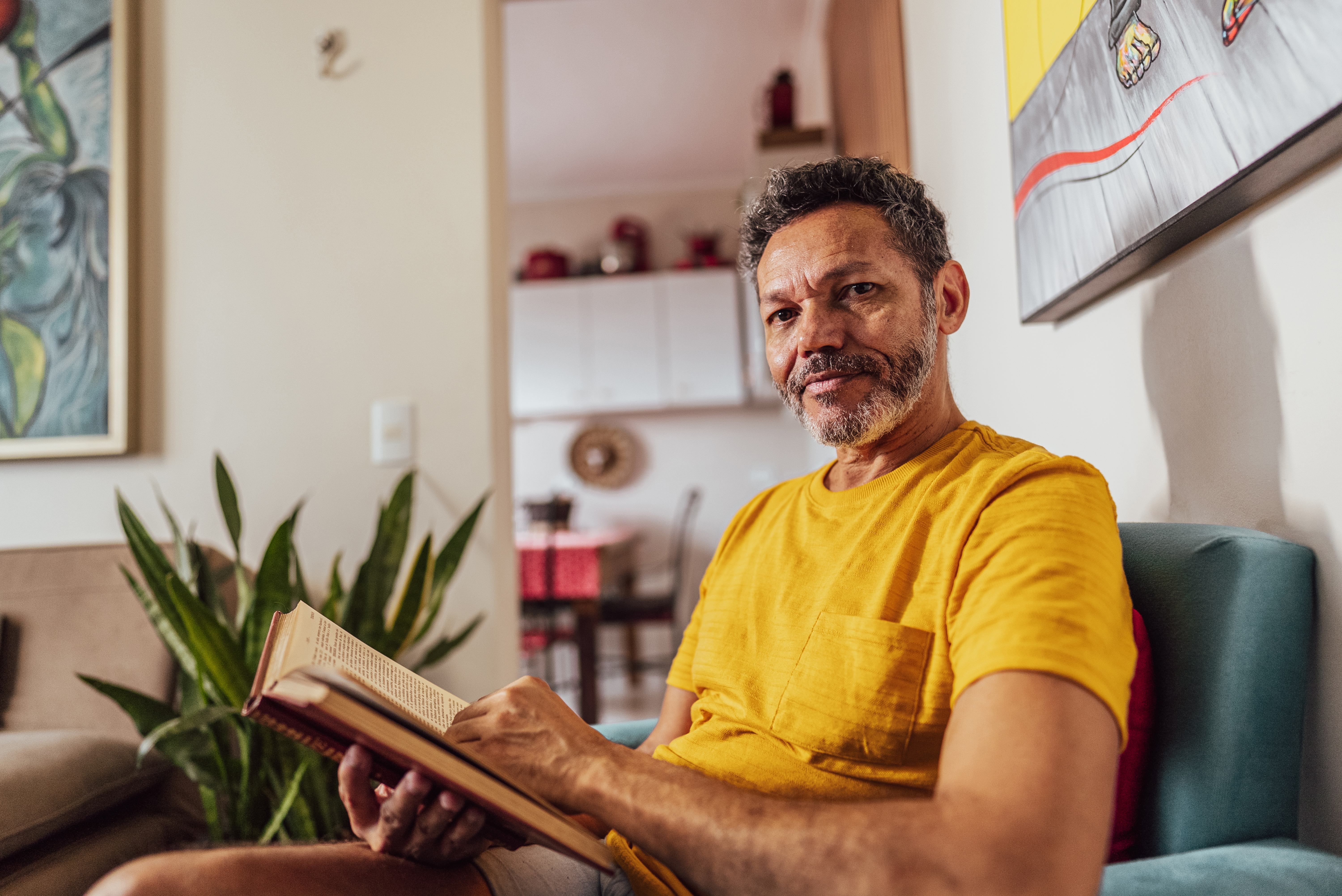 Mann i uformelle omgivelser, sitter i en sofa, leser en bok og ser på kameraet