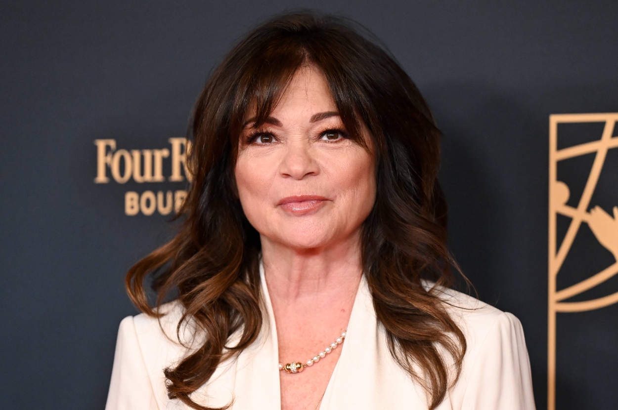 A person stands on the red carpet wearing a white suit with a pearl necklace, smiling for the camera