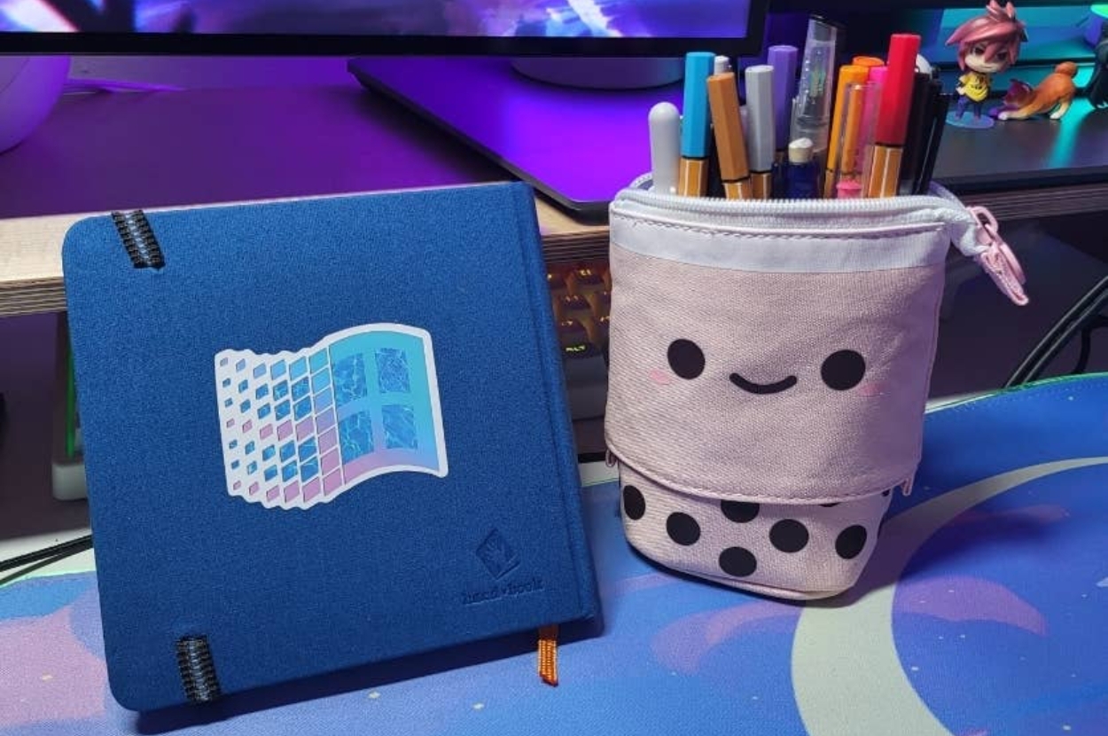 Smiling pen holder with various pens beside a notebook with a retro design on a desk setup