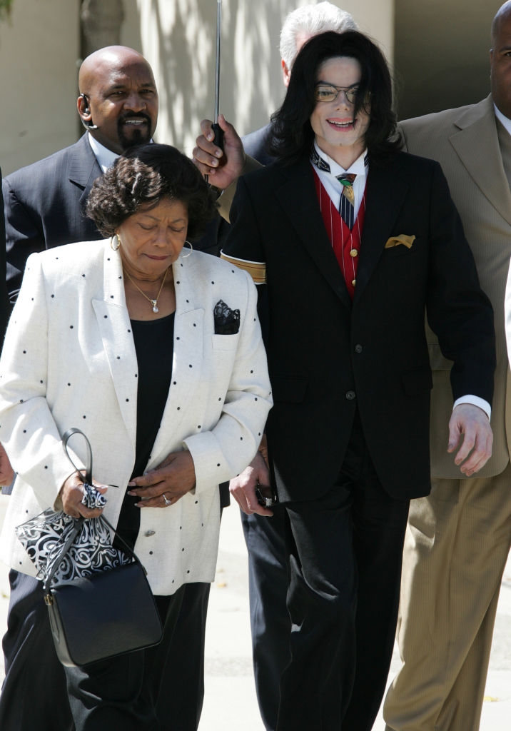 A idiosyncratic successful a stylish overgarment walks with an older pistillate successful a polka dot jacket, accompanied by information personnel