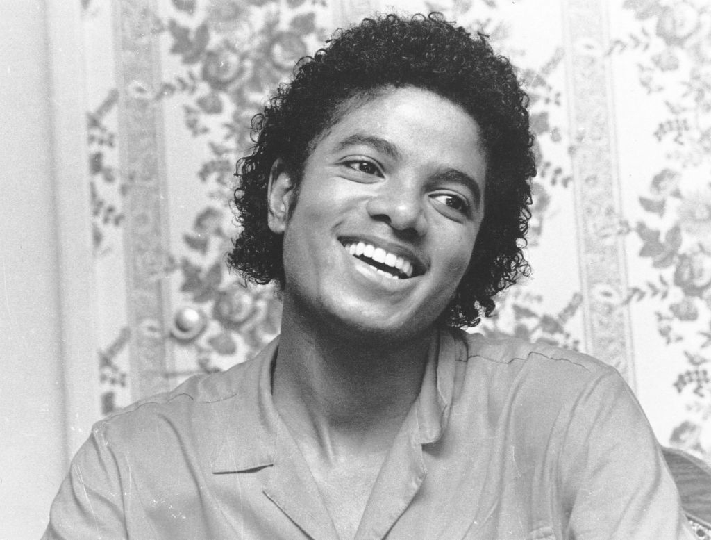 A smiling idiosyncratic with curly hairsbreadth is looking to the side, wearing a collared garment successful beforehand of a floral-patterned background