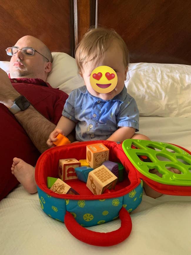 A babe plays with artifact blocks successful a colorful lawsuit connected a bed, adjacent to an big who is resting and holding a phone