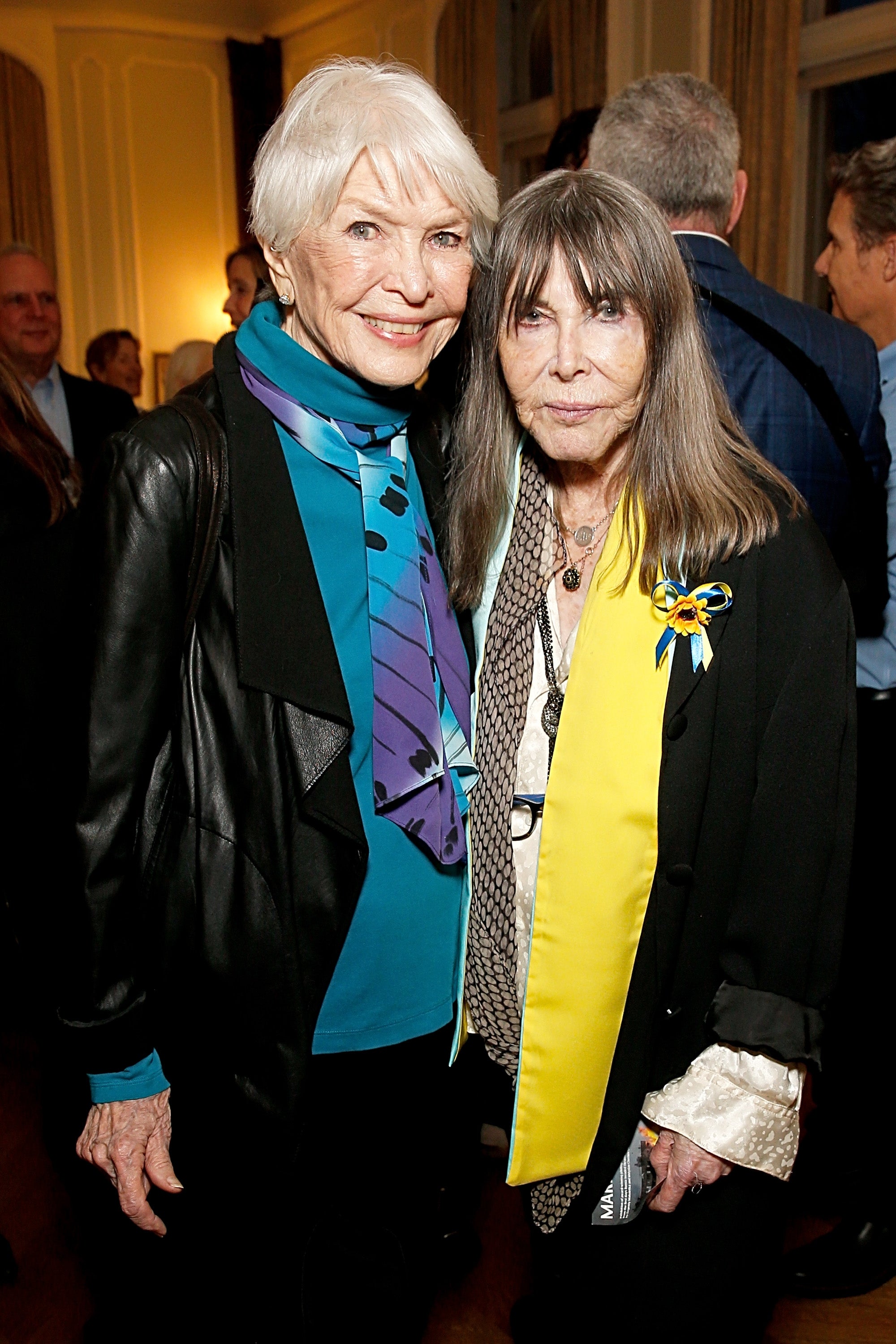 Ellen and Lee smiling at an art gallery, one wearing a leather jacket with a colorful scarf, the other in a black outfit with a yellow and blue ribbon