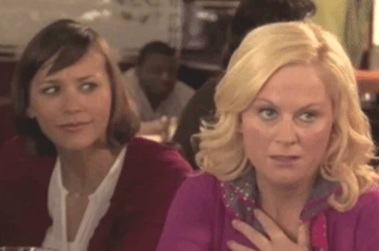 Two women sitting at a table, one looking skeptical and the other appearing surprised, hand on chest