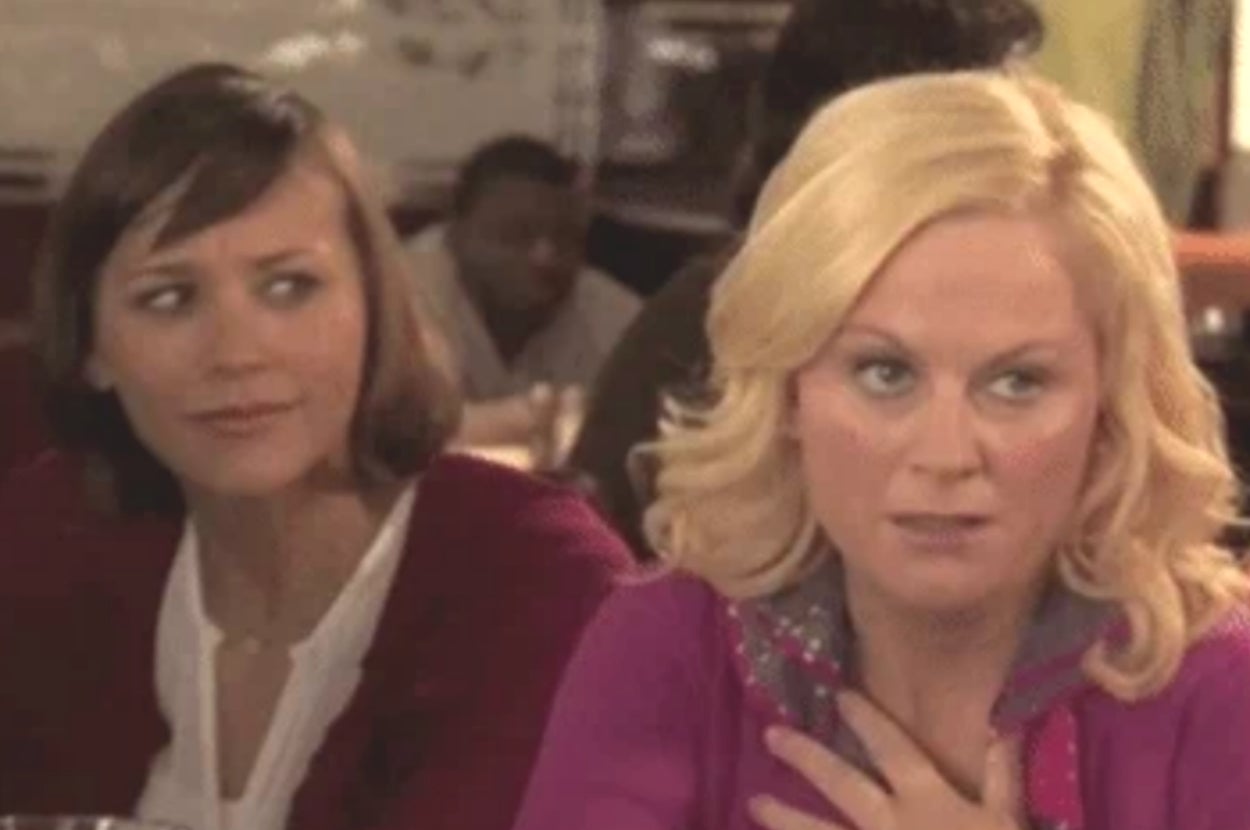 Two women sitting at a table, one looking skeptical and the other appearing surprised, hand on chest