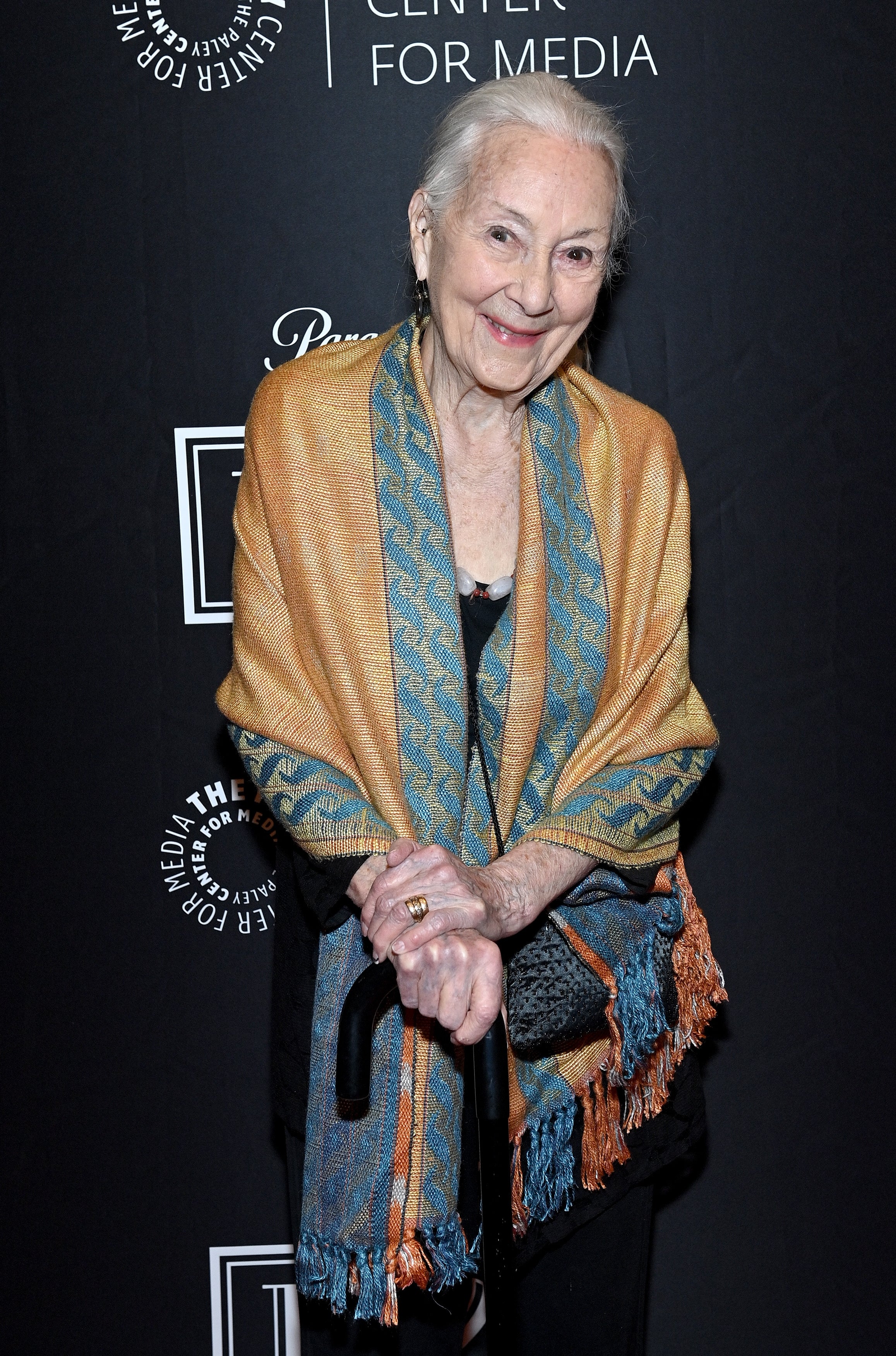 An elderly woman stands on the red carpet, smiling, wearing a shawl over her shoulders and holding a cane, at The Paley Center for Media event