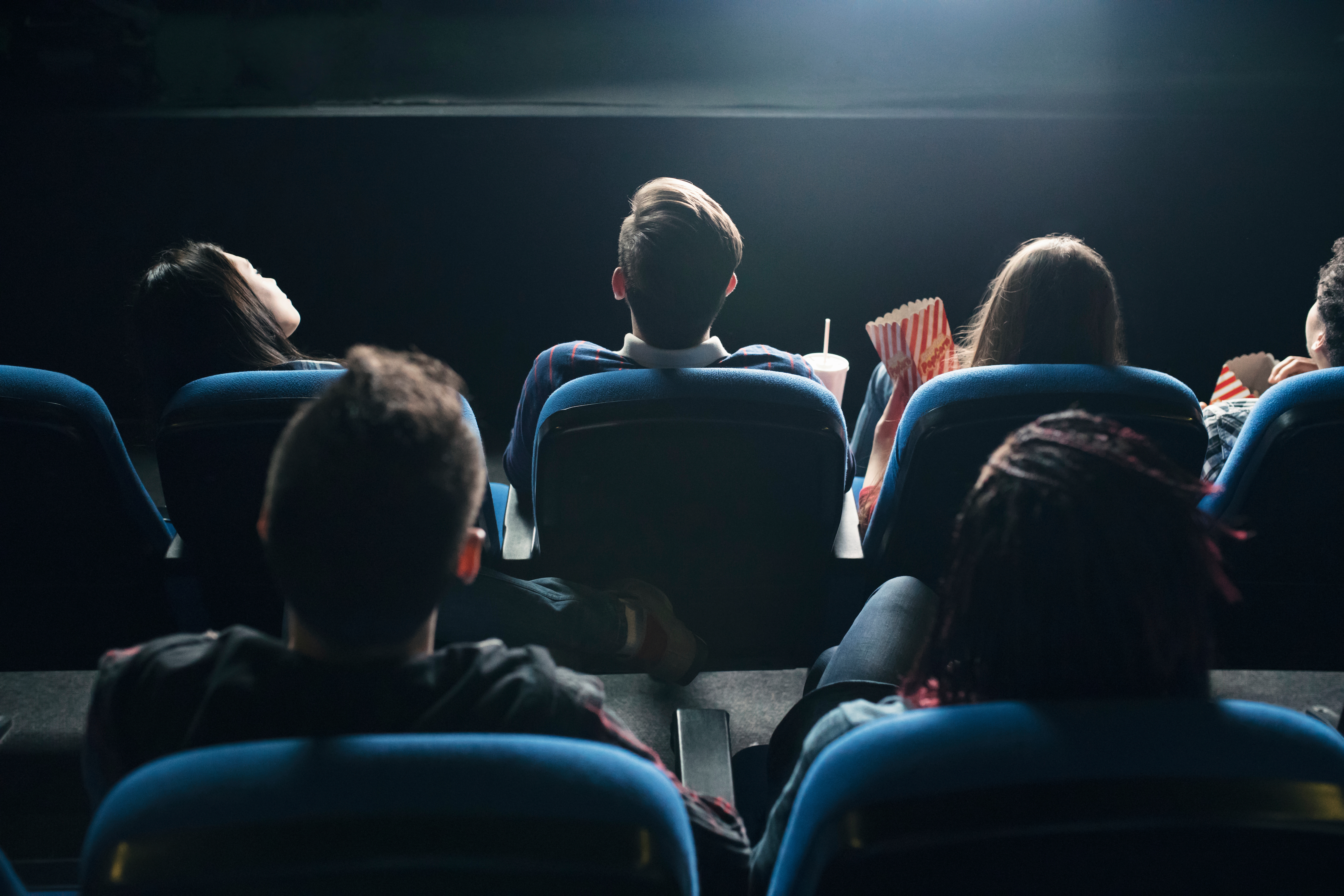 People sitting in a movie theater, seen from behind, holding drinks and popcorn, watching a film on the big screen