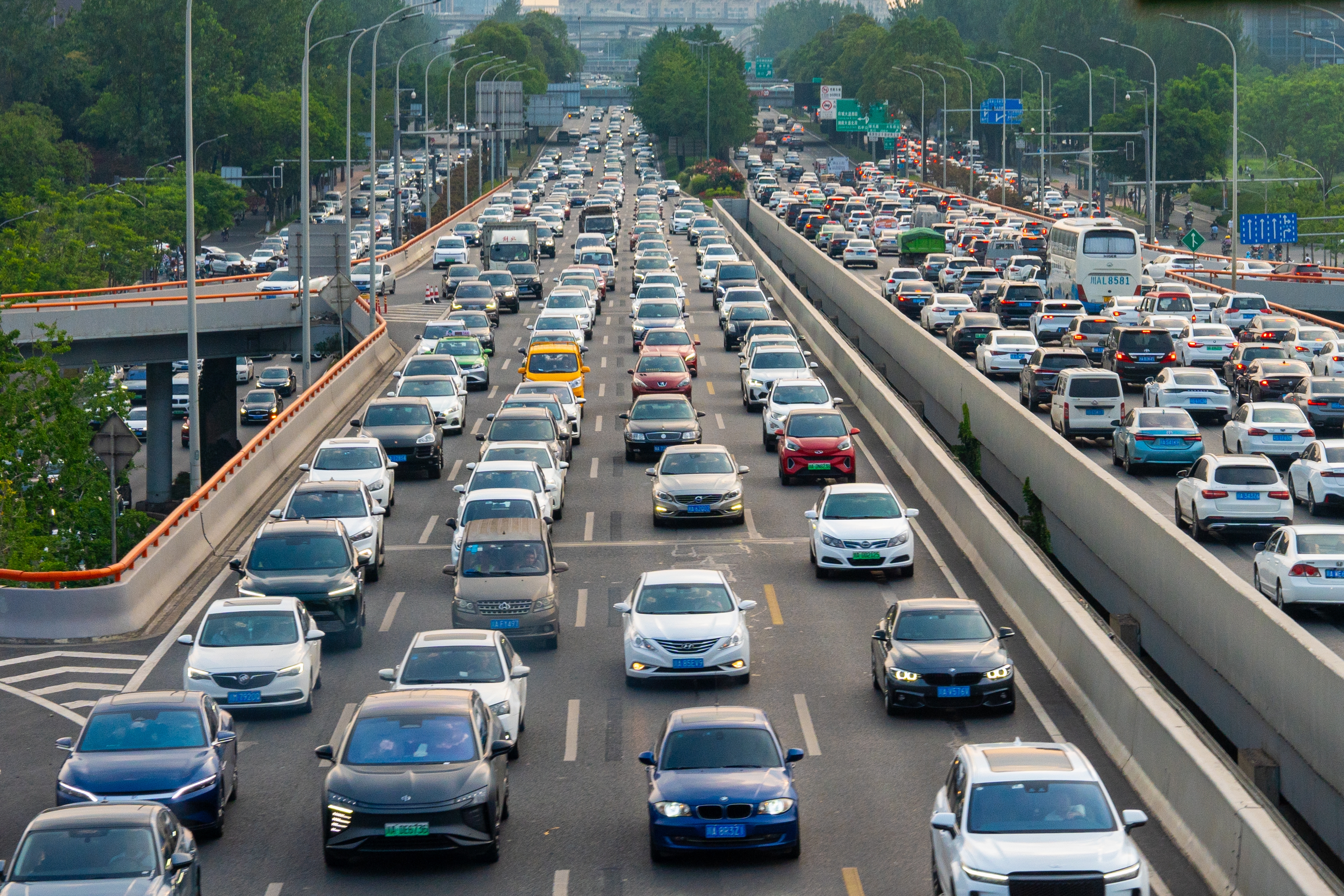 Traffic jam on a multi-lane highway with cars and buses moving slowly, surrounded by trees and overpasses in an urban setting