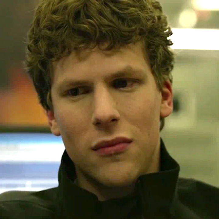 Person with short curly hair looks down with a thoughtful expression, wearing a dark jacket indoors