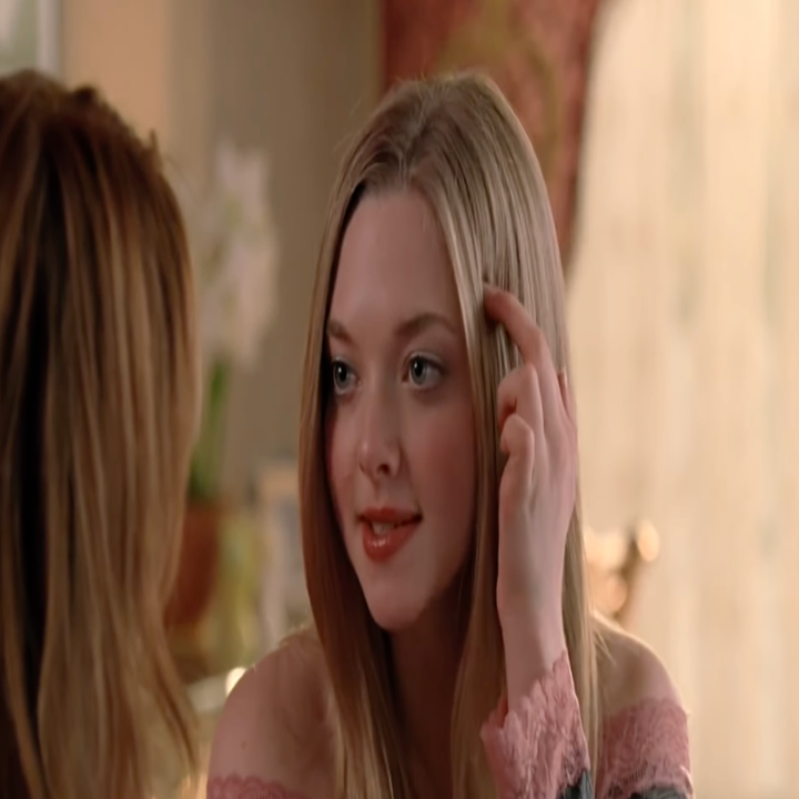 A person with long hair is sitting indoors, gesturing with one hand. They are speaking to someone off-camera