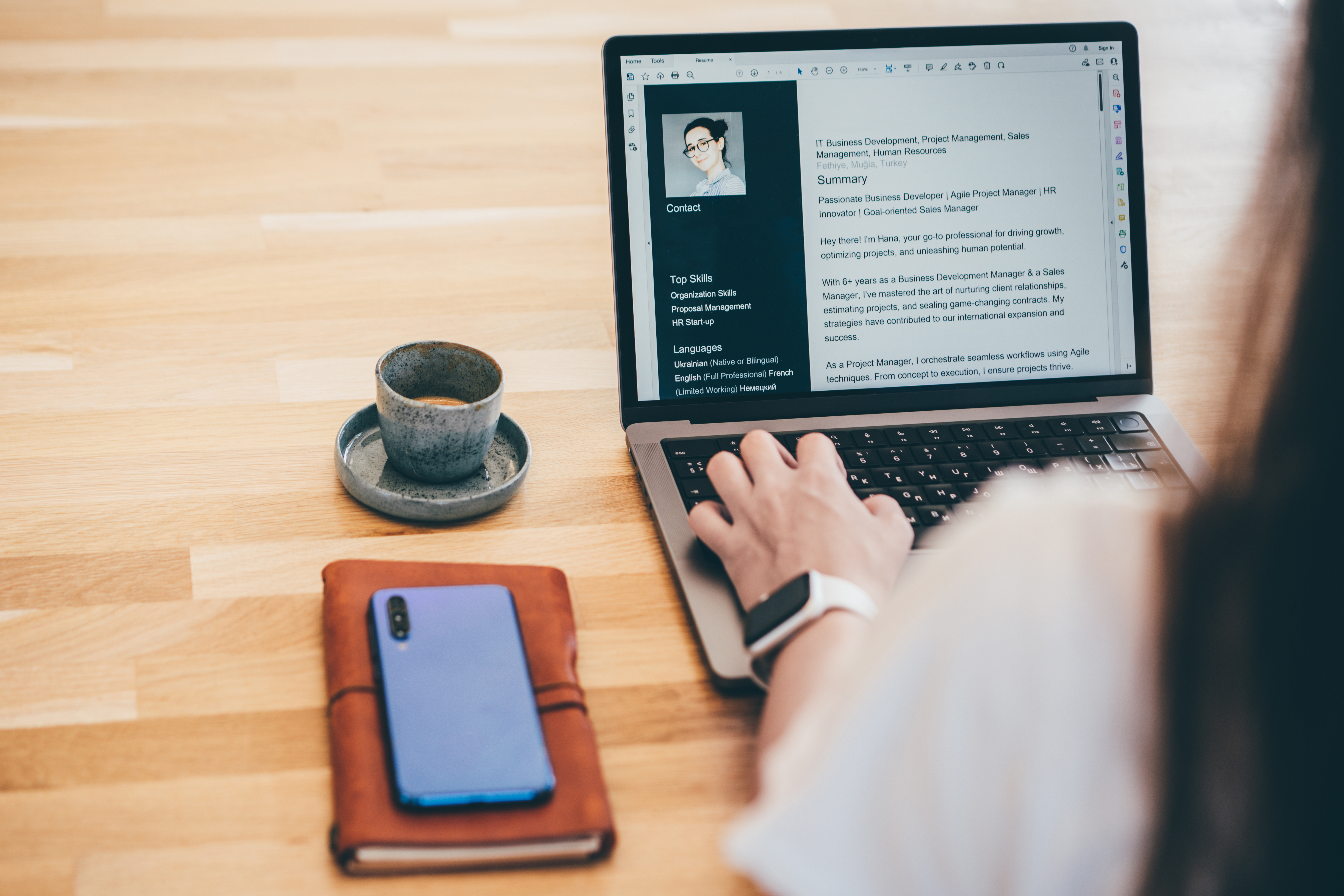Person typing on a laptop with a resume on screen, next to a coffee cup, notebook, and smartphone on a wooden desk
