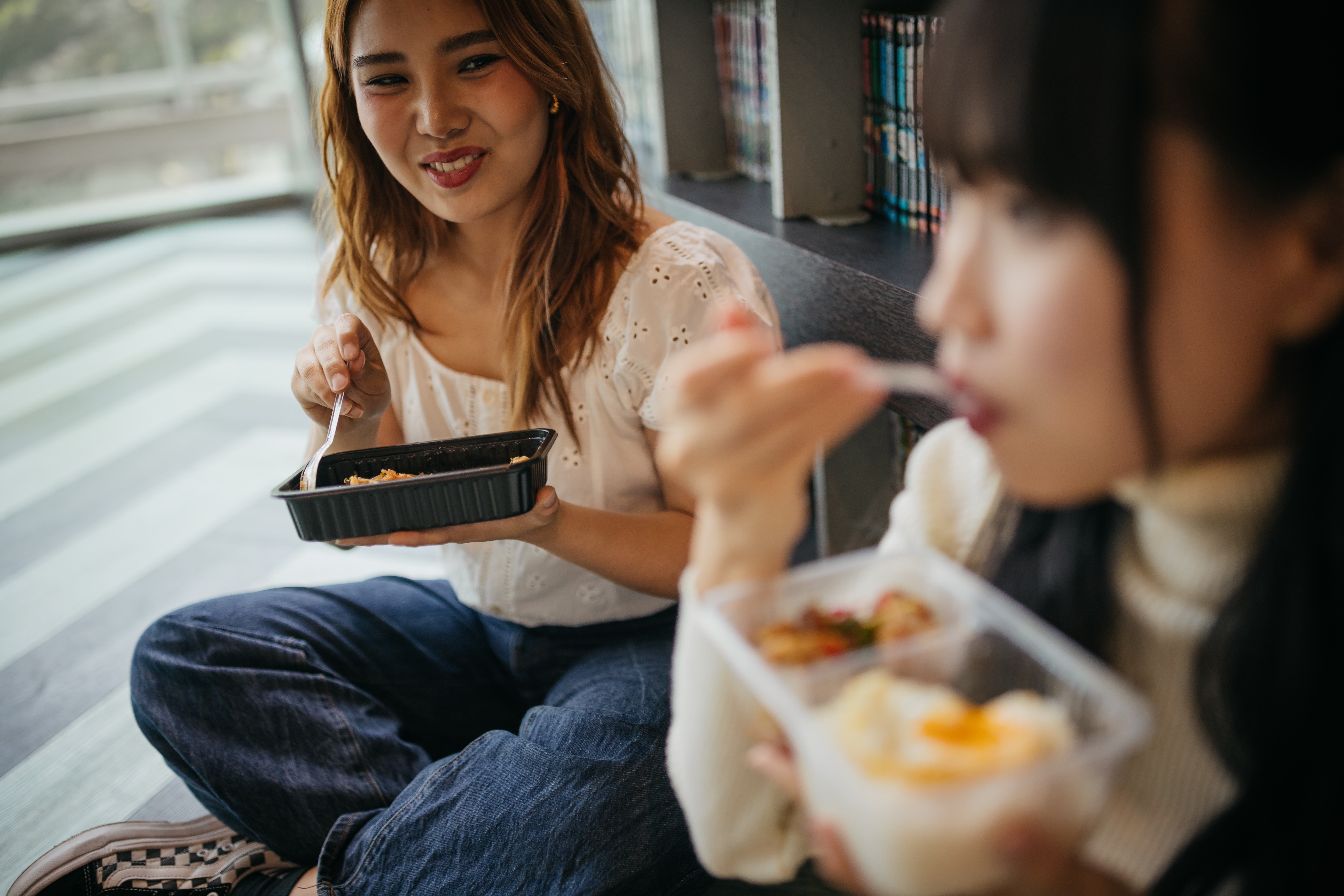 Two people sit together, smiling and eating from food containers, suggesting a casual, friendly meal setting indoors