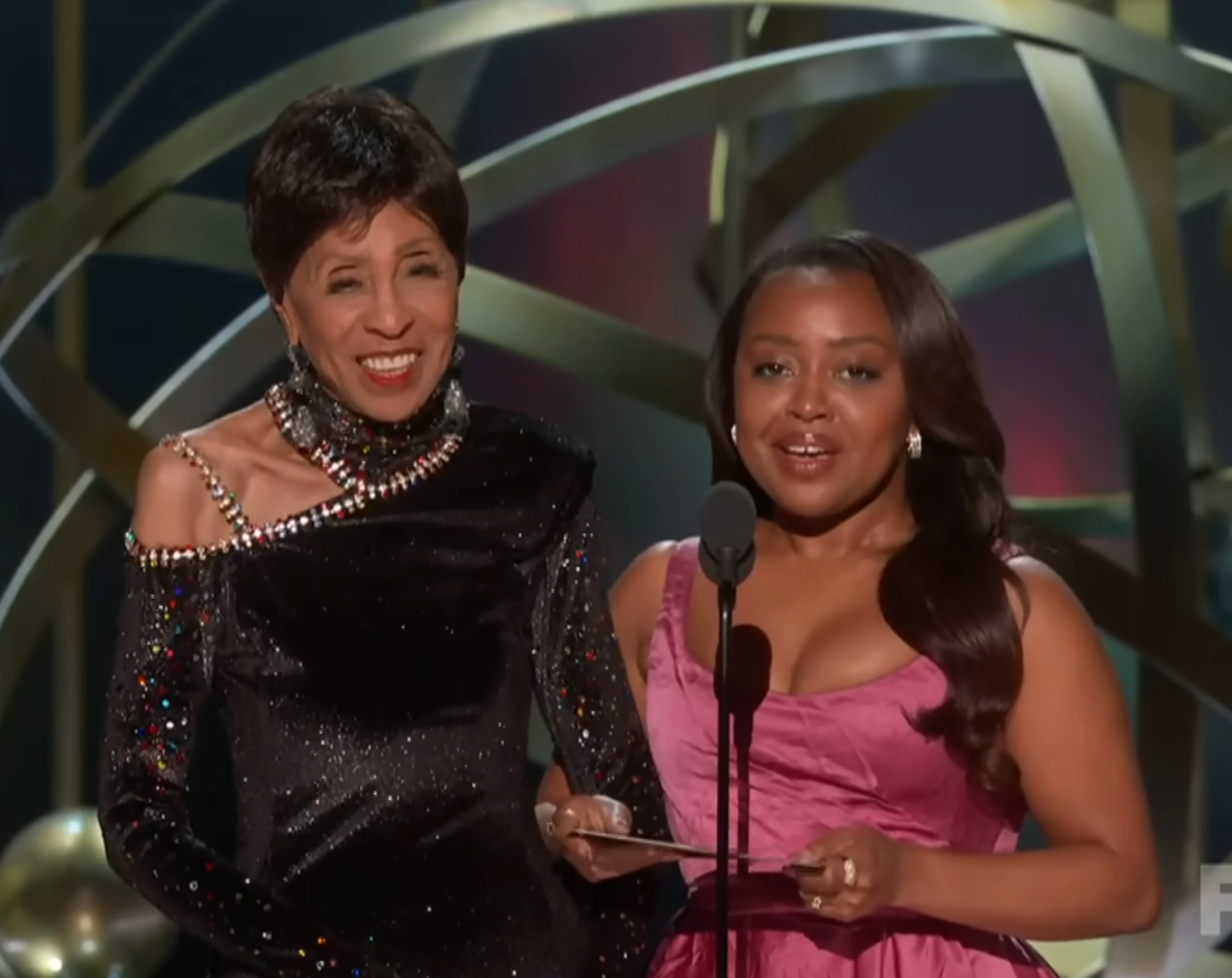 Two women on stage presenting an award. One wears a bejeweled black gown; the other is in a stylish sleeveless gown. They hold an envelope