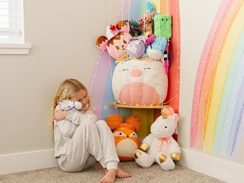Child sitting with a plush artifact  collection, wearing a cozy outfit, adjacent  to a partition  with a rainbow design