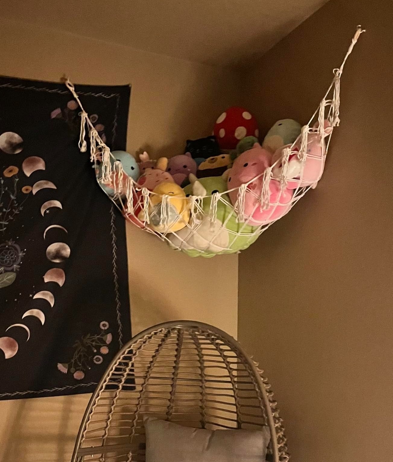 Hammock holding plush toys against a wall, adjacent   a tapestry with satellite  phases, supra  a wicker seat  with a cushion