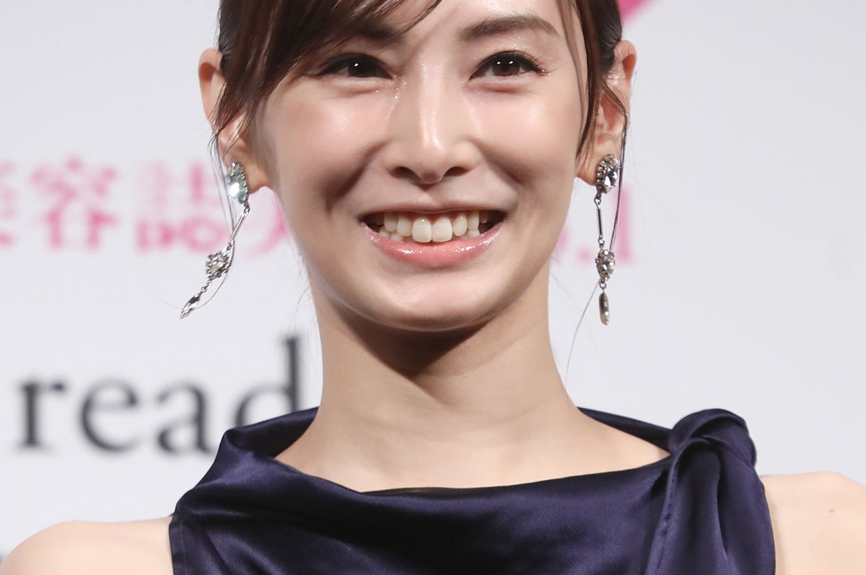 有名人がイベントで光沢のあるドレスを着て微笑む。後ろに広告用の背景がある。