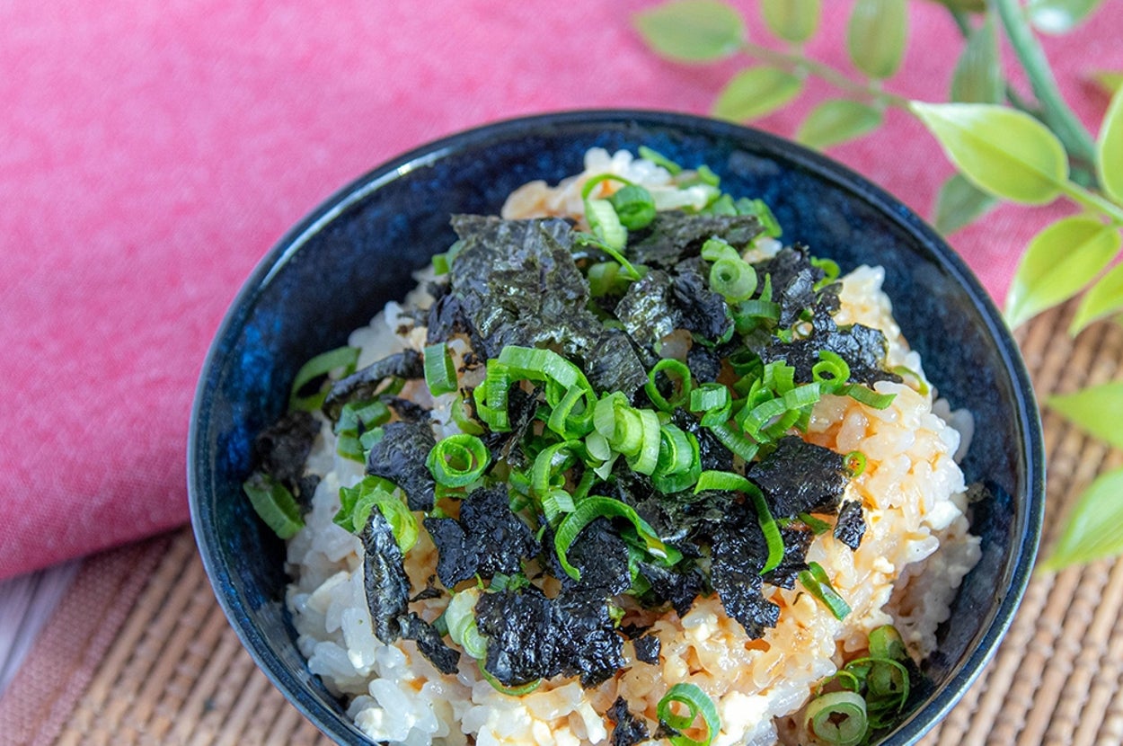 のりとねぎがのったライスボウルが竹のマットに置かれている。横には赤い布と緑の装飾がある。