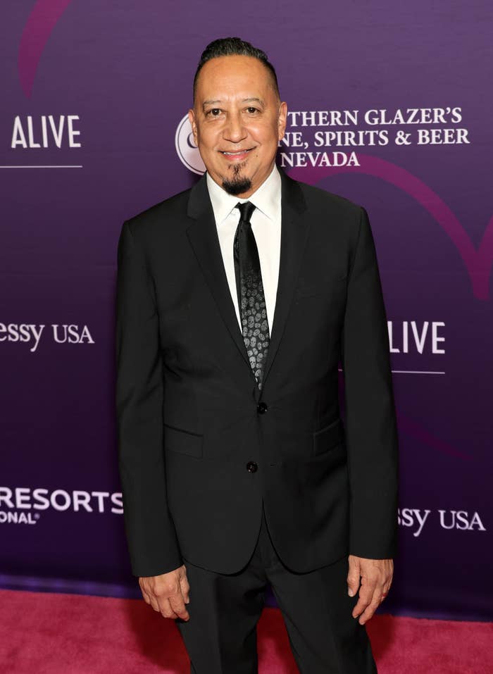 A person in a black suit with a tie poses on a red carpet at an event, with a backdrop of sponsor logos