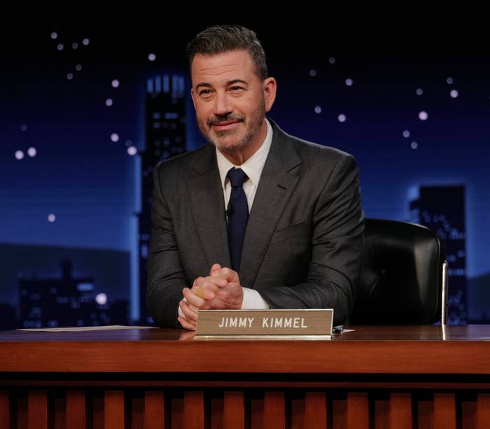 Person in a dark suit and tie sits at a desk with a nameplate, appearing to host a late-night talk show under a city skyline backdrop