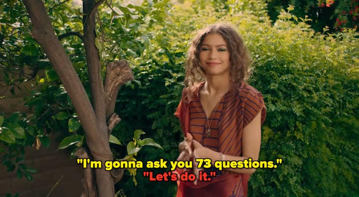 Woman with curly hair in a striped blouse and skirt stands by a tree in a garden, smiling subtly