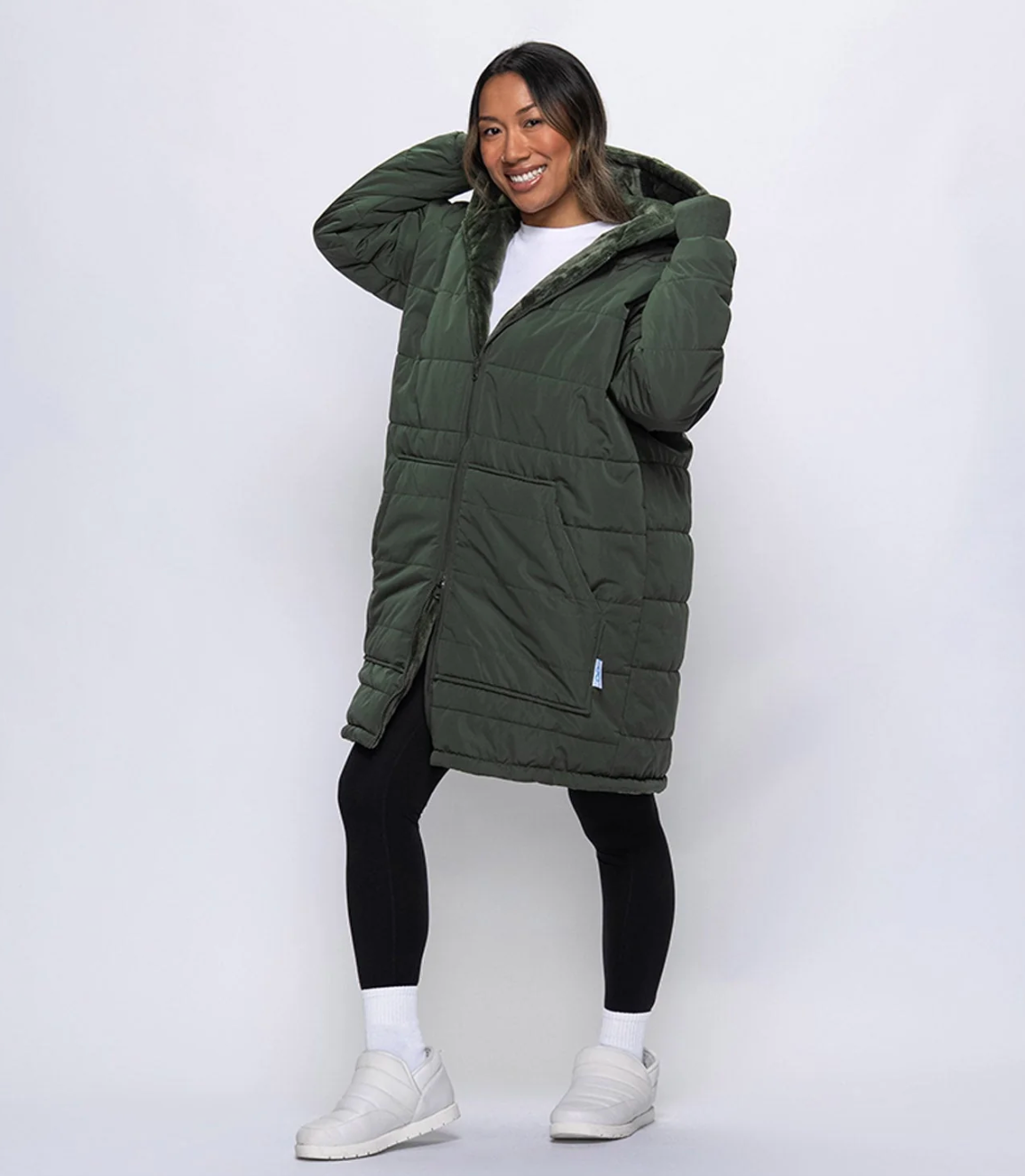 Person smiling, wearing a agelong quilted overgarment with a hood, achromatic leggings, and achromatic sneakers, striking a casual pose