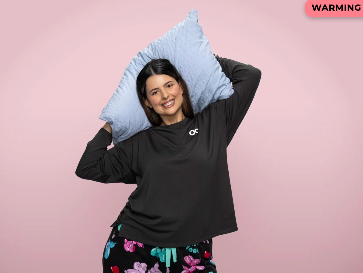 Person smiling, holding a pillow down their head, wearing a casual sweatshirt and printed pants. Perfect for a cozy, relaxed buying theme