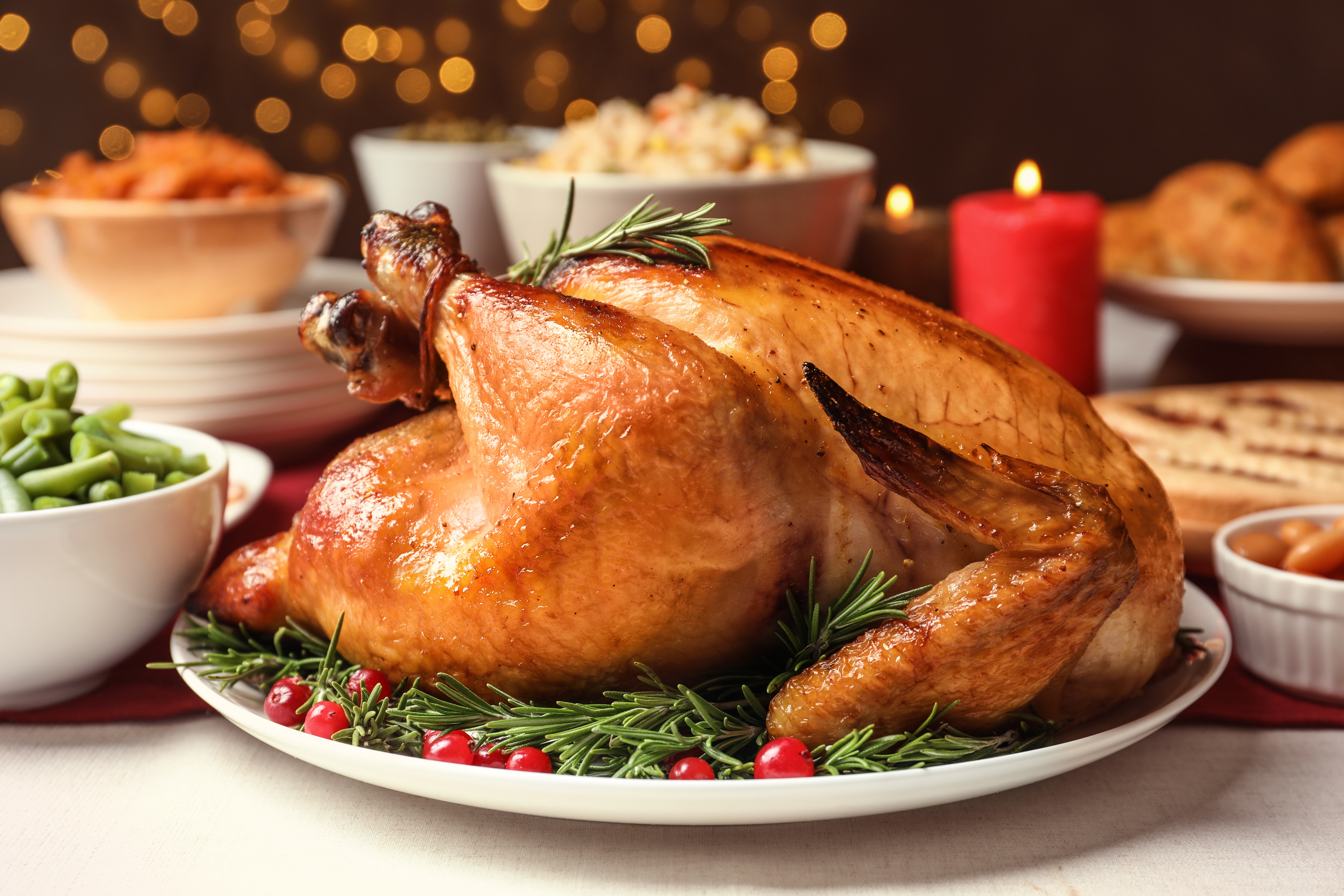 A roasted turkey garnished with rosemary and cranberries on a festive table. Background includes bowls of sides and a lit red candle
