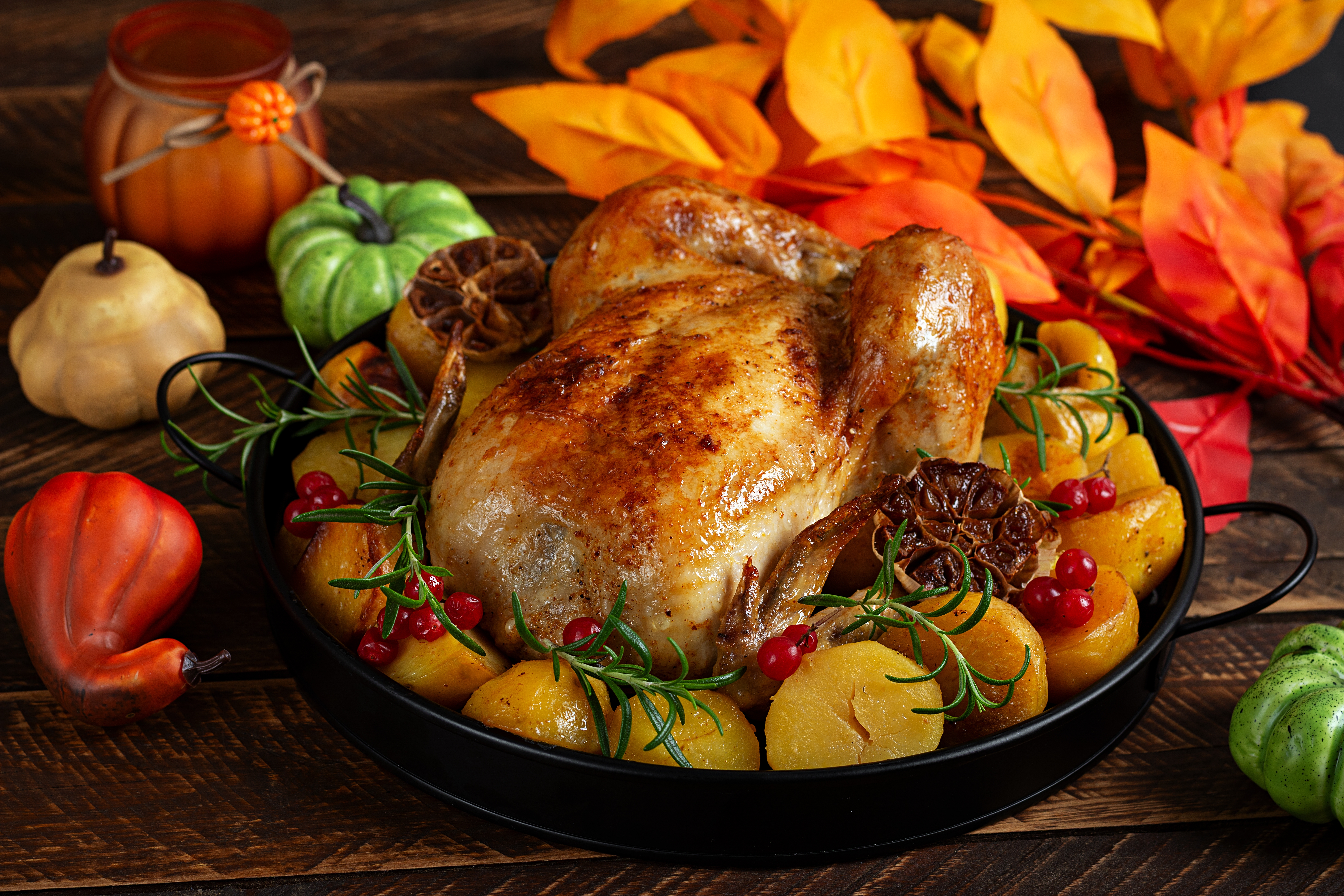 Roasted chicken surrounded by potatoes, rosemary, red berries, and decorative gourds on a wooden table with autumn leaves