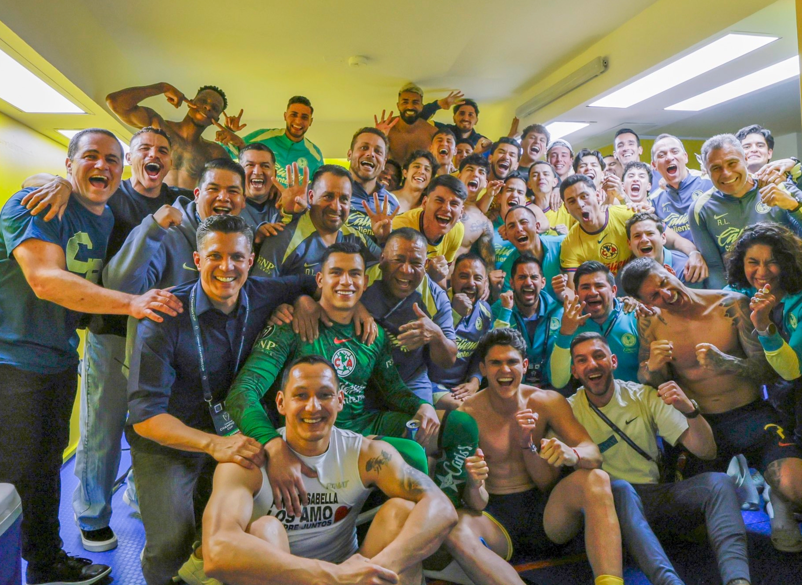 Group of excited soccer players and staff celebrating in a locker room, some in jerseys, others in casual wear, all showing joyful expressions