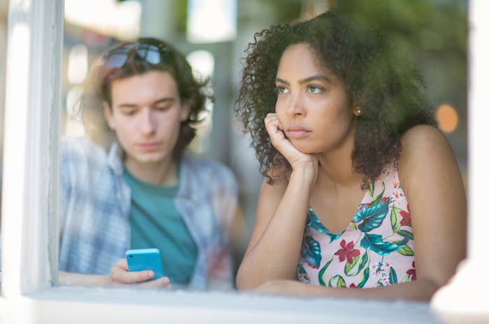 Two radical   are sitting by a window. One looks astatine  a phone, portion    the different   stares outside, appearing thoughtful