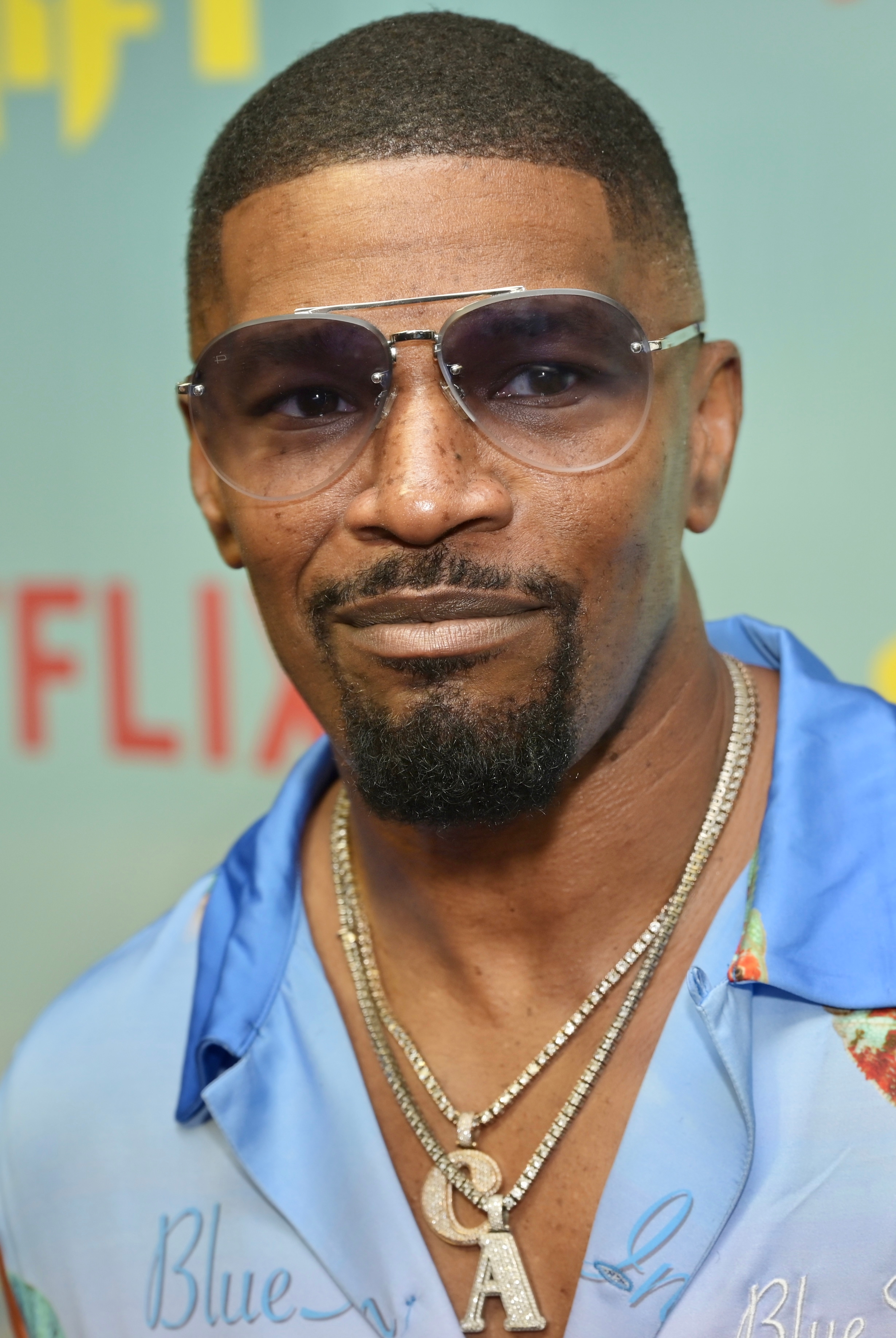 Man connected reddish carpet wearing sunglasses, a bluish patterned shirt, and layered necklaces with a salient missive "A" pendant
