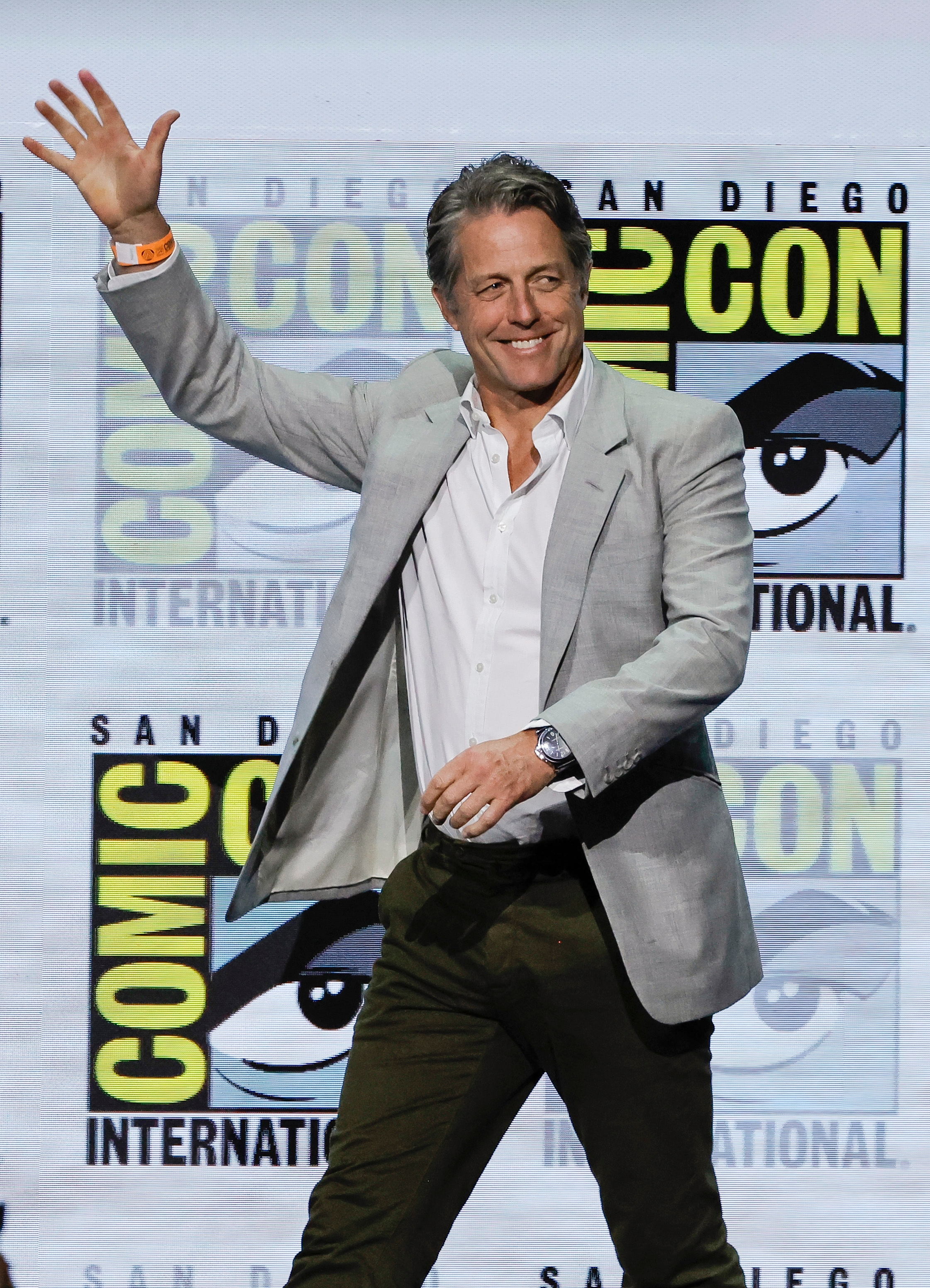 Man successful a casual suit waves astatine Comic-Con event, smiling with a backdrop of Comic-Con logos