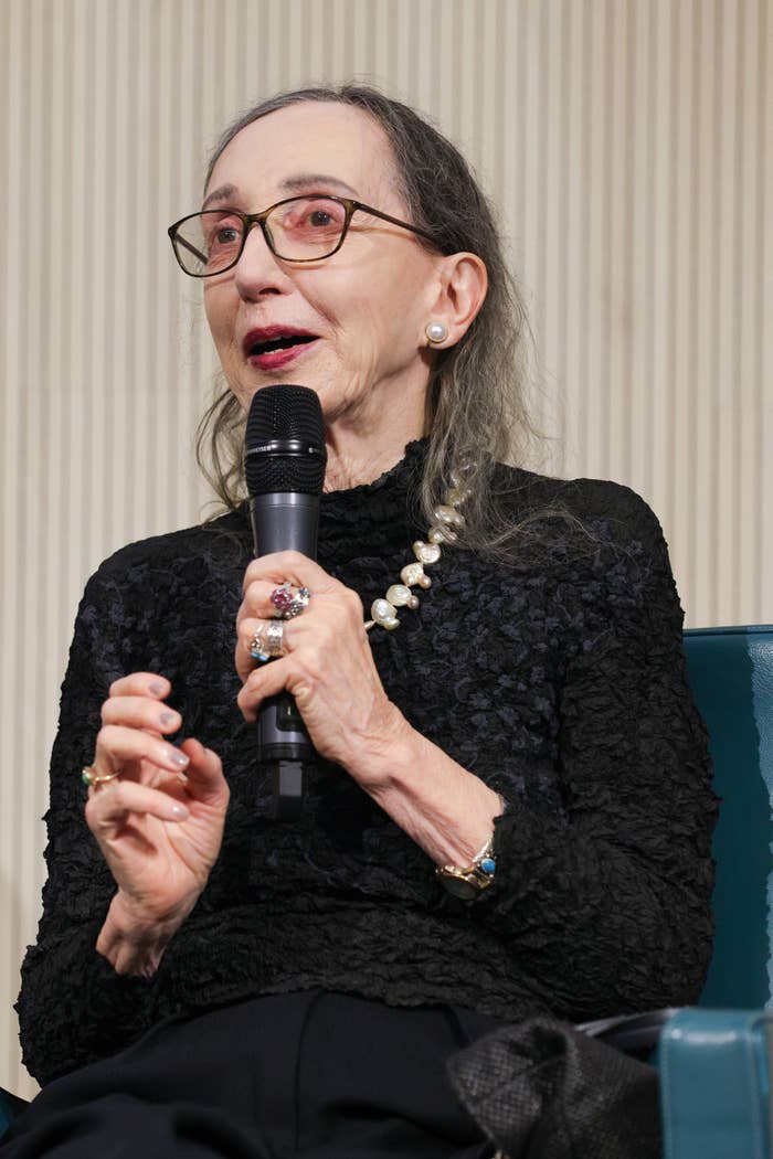 Person with glasses, wearing a textured black top and pearl necklace, speaks into a microphone at an event