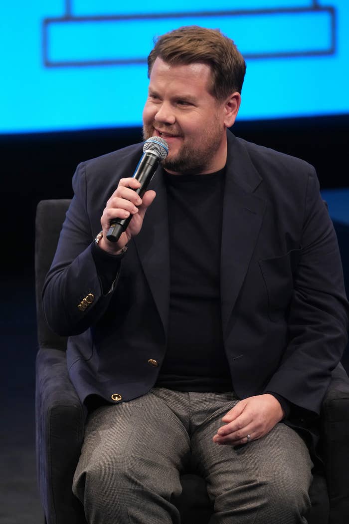 Person seated connected  stage, wearing a acheronian  blazer and pants, holding a microphone