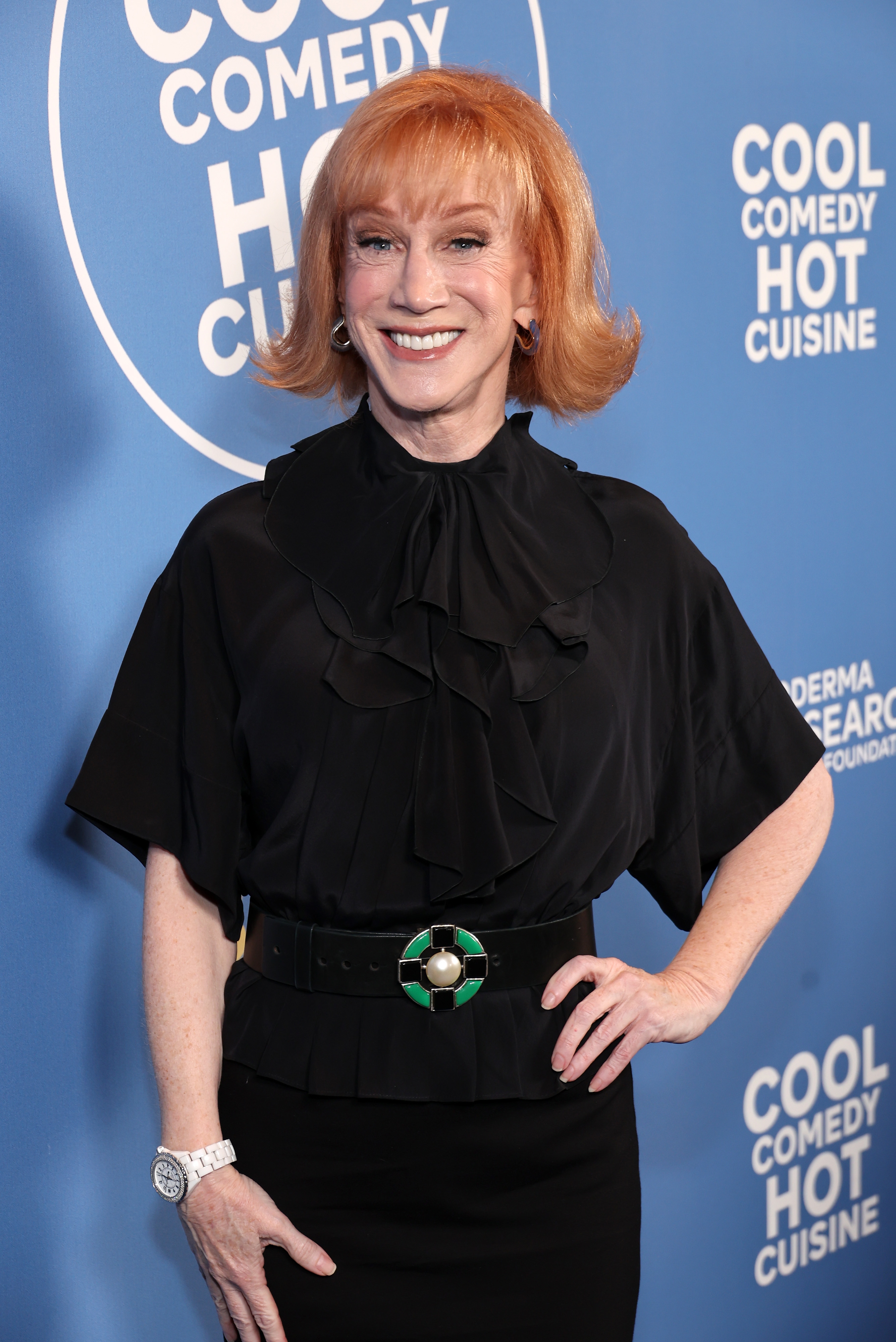 Person smiling connected the reddish carpet successful a achromatic outfit with a ruffled top, attending a "Cool Comedy Hot Cuisine" event