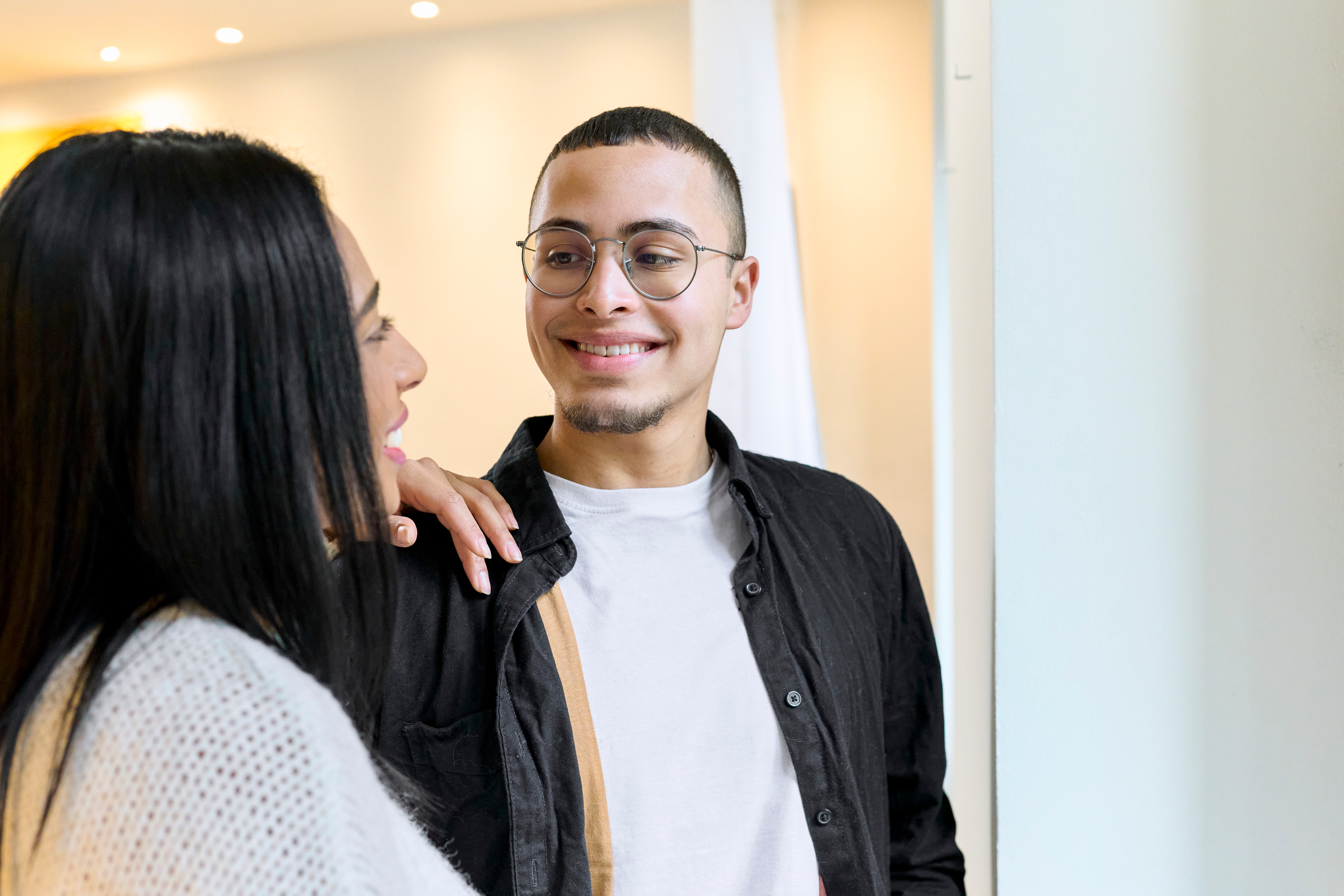 Two radical smiling astatine each different warmly indoors, 1 wearing glasses and a achromatic garment implicit a achromatic tee, the different successful a cozy sweater