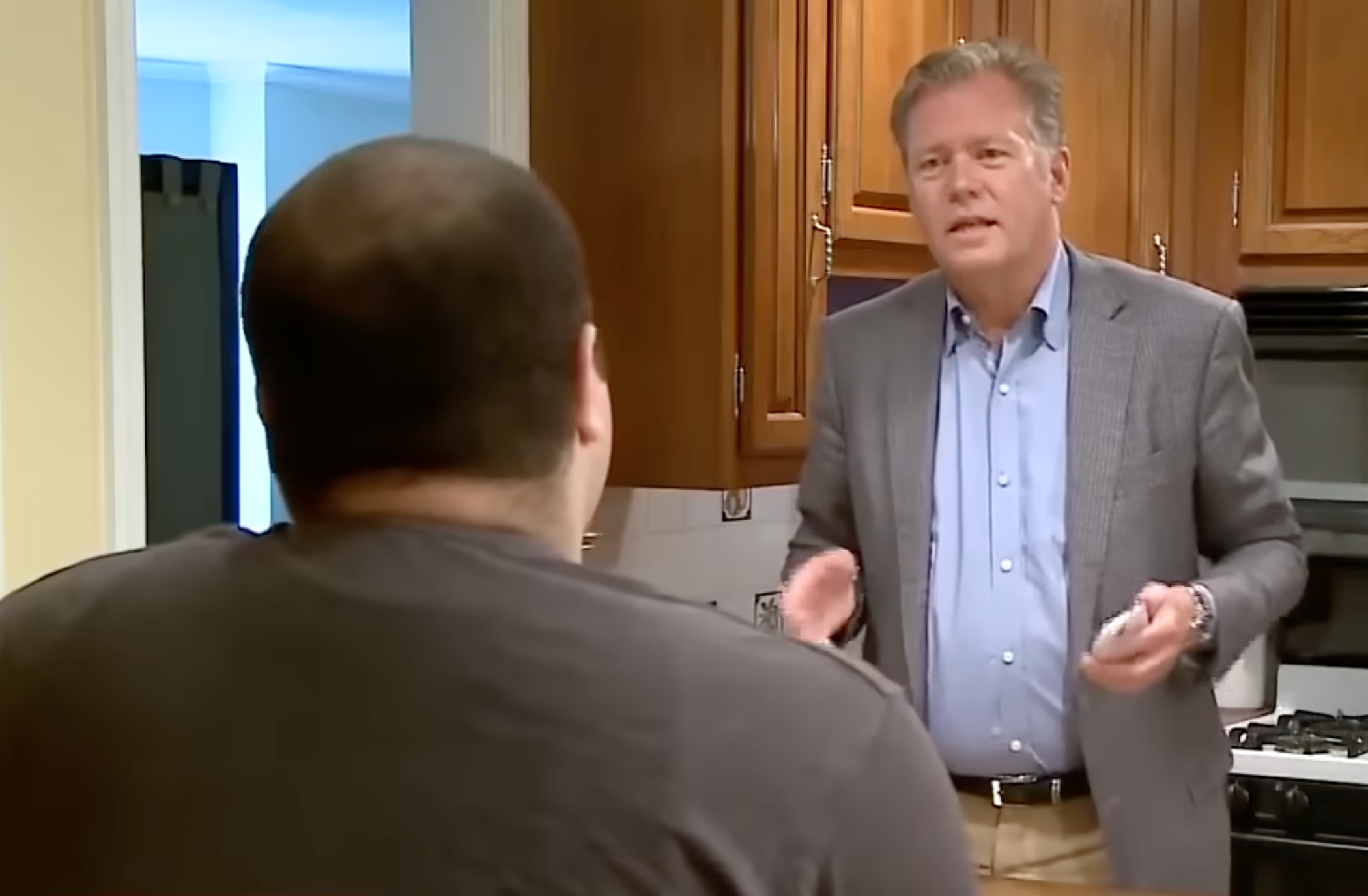 Two men are successful  a kitchen. One is seated with his backmost  to the camera; the different   is standing, wearing a suit, and appears to beryllium  speaking