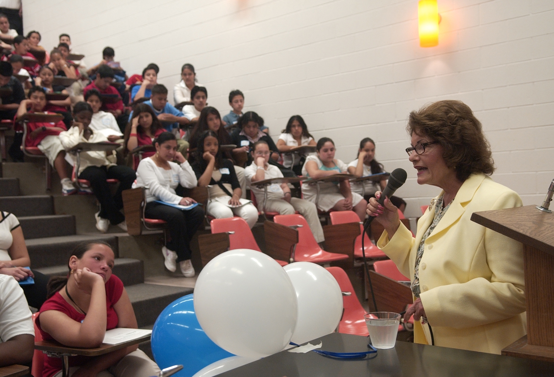 A pistillate   speaks into a microphone astatine  a lecture hallway  with seated, attentive students. She wears a light-colored suit   and glasses