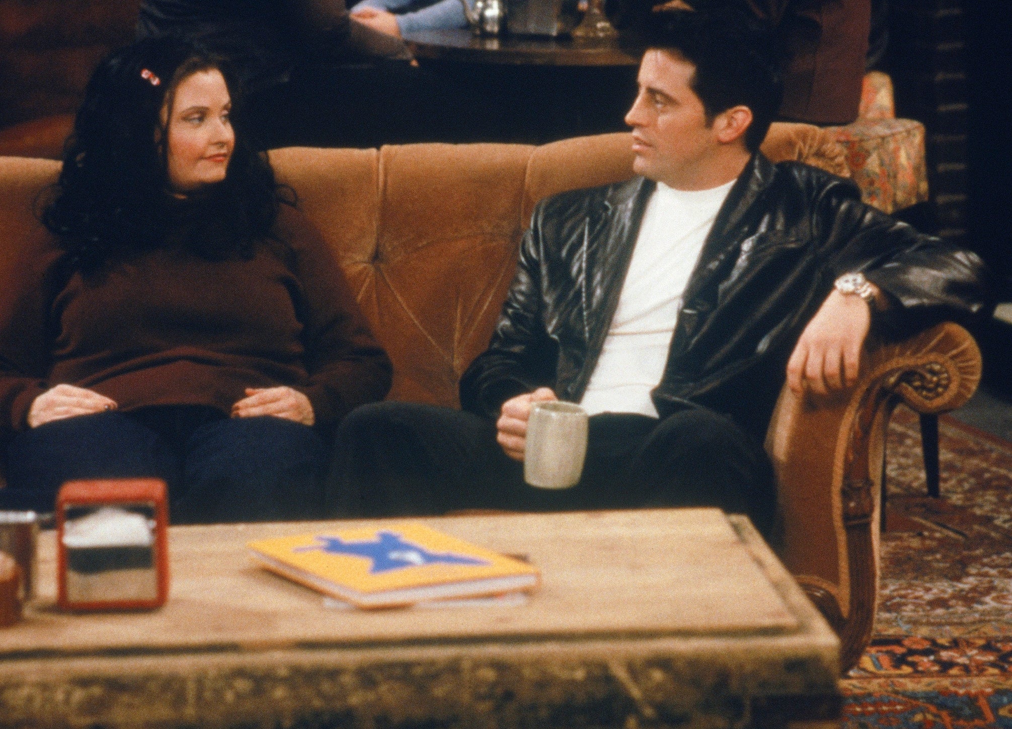 Two radical   beryllium   connected  a sofa  successful  a lively cafe setting, conversing and holding cups, with others chatting and lasting  successful  the background