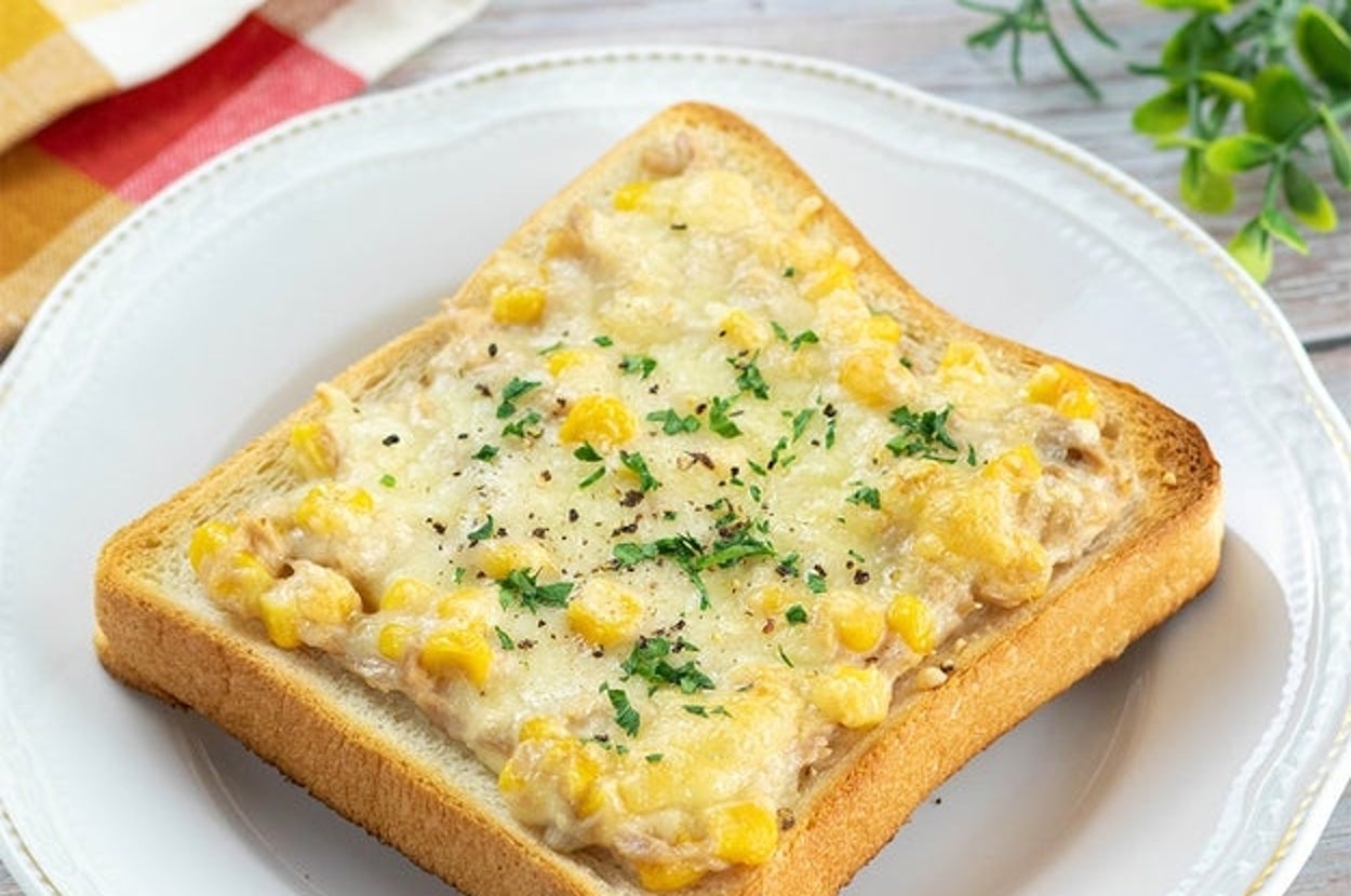 トーストにコーン、チーズ、パセリが載った朝食用の料理。背景に布ナプキンと観葉植物があります。