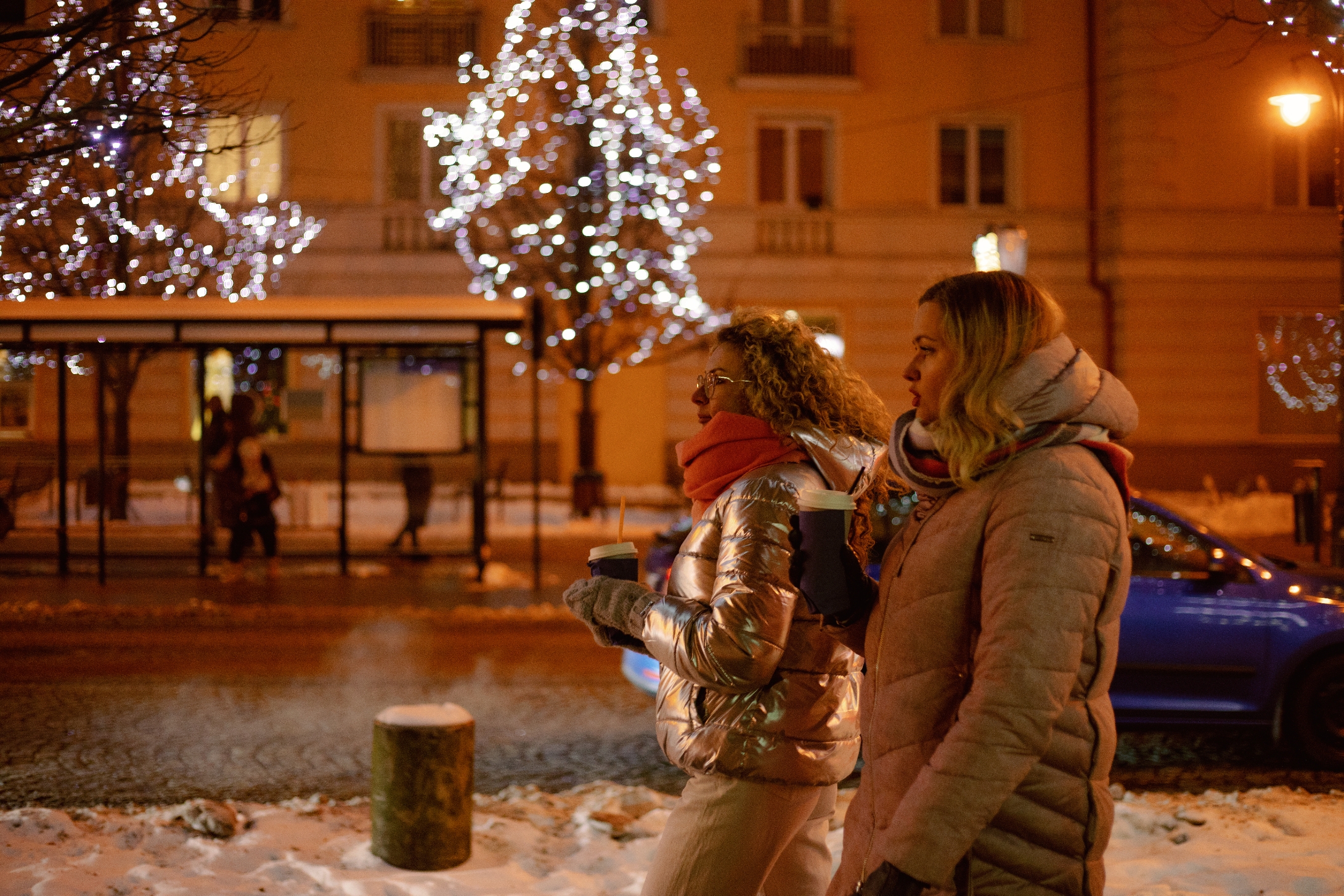 Two radical   successful  wintertime  coats clasp  cups, walking past   a snowy thoroughfare  with illuminated trees and buildings successful  the background