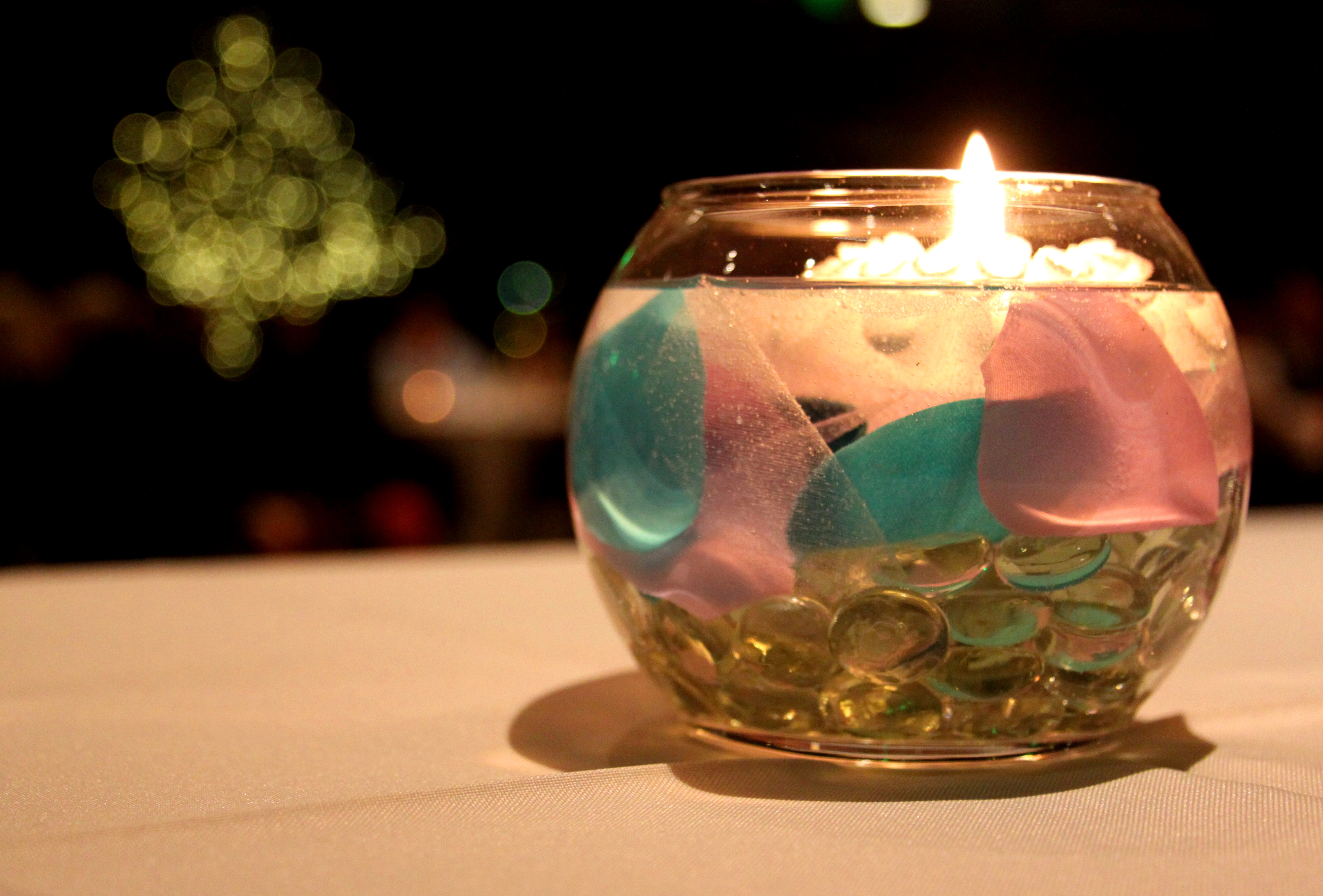 Glass vessel  centerpiece with floating candle, pinkish  and bluish  petals, and solid  beads. Blurred lights successful  the background