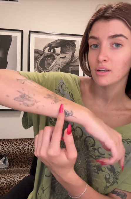 Person pointing astatine  tattoos connected  their forearm portion    seated indoors. They&#x27;re wearing a casual patterned apical  with framed photos successful  the background